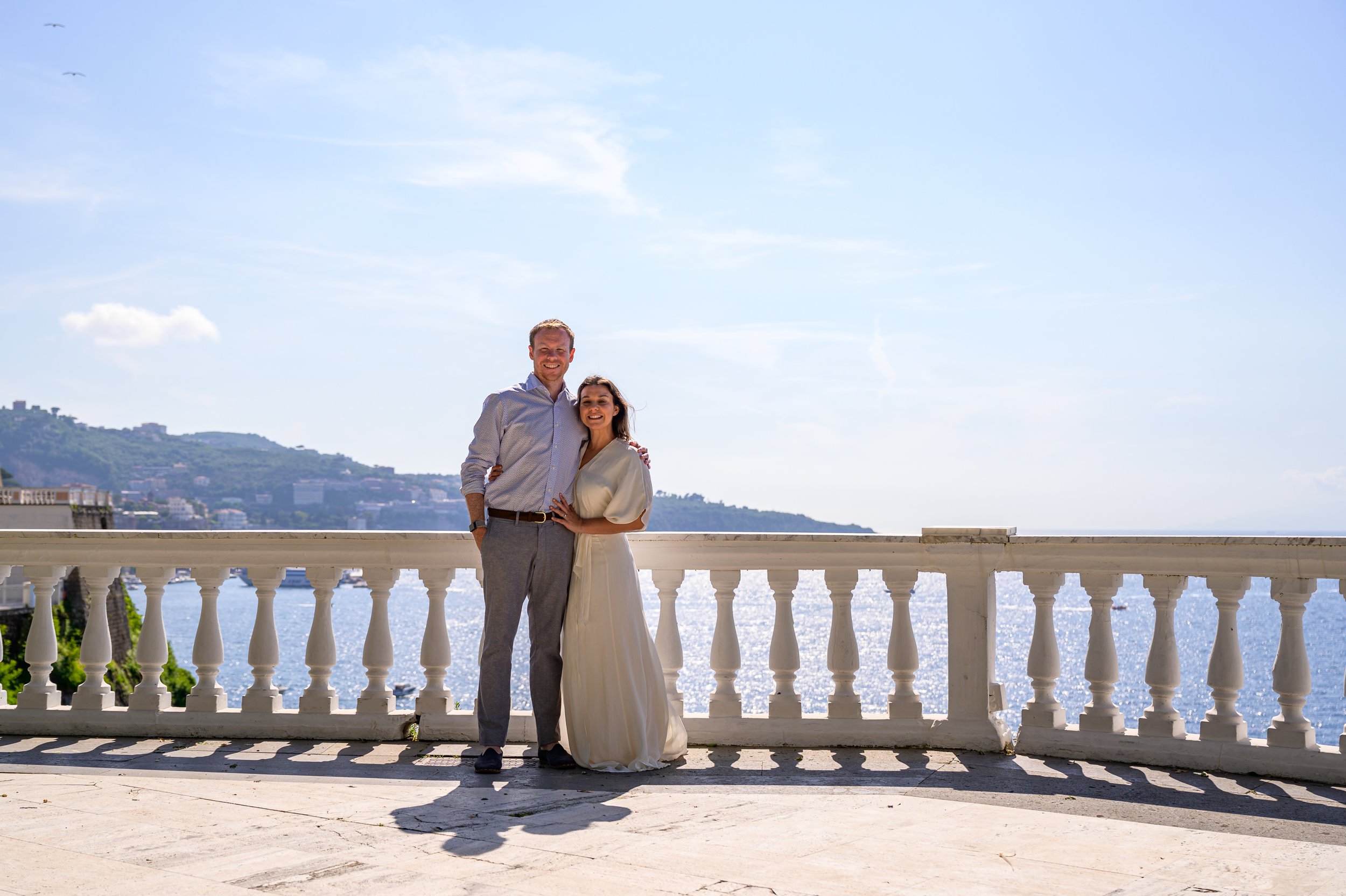 A smiling couple in formal attire stands on a balcony by a body of water, with hills and buildings in the distance under a clear blue sky.