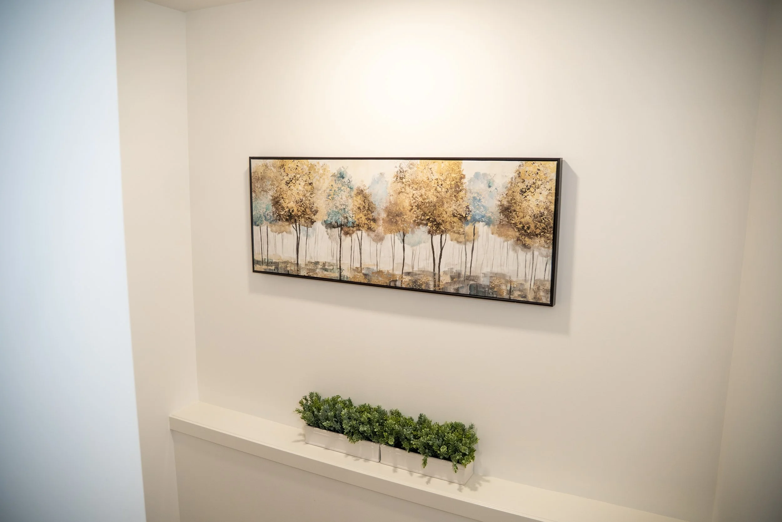 Interior wall with a framed painting of trees in autumn colors and a potted green plant on a white ledge.