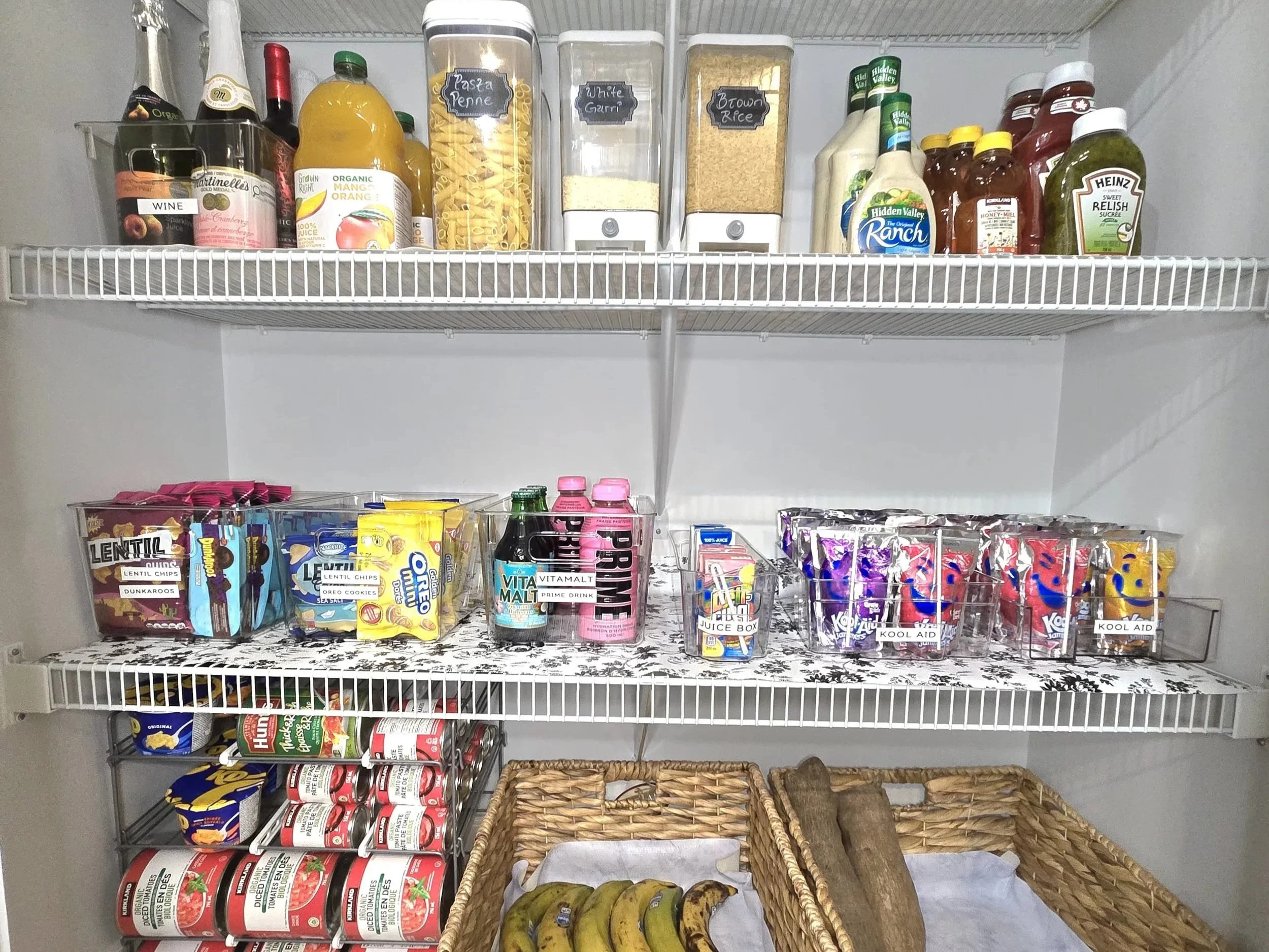 Open pantry with three shelves filled with various food and beverage items, including canned goods, snacks, drinks, condiments, and dry ingredients, along with baskets containing bananas and a log.