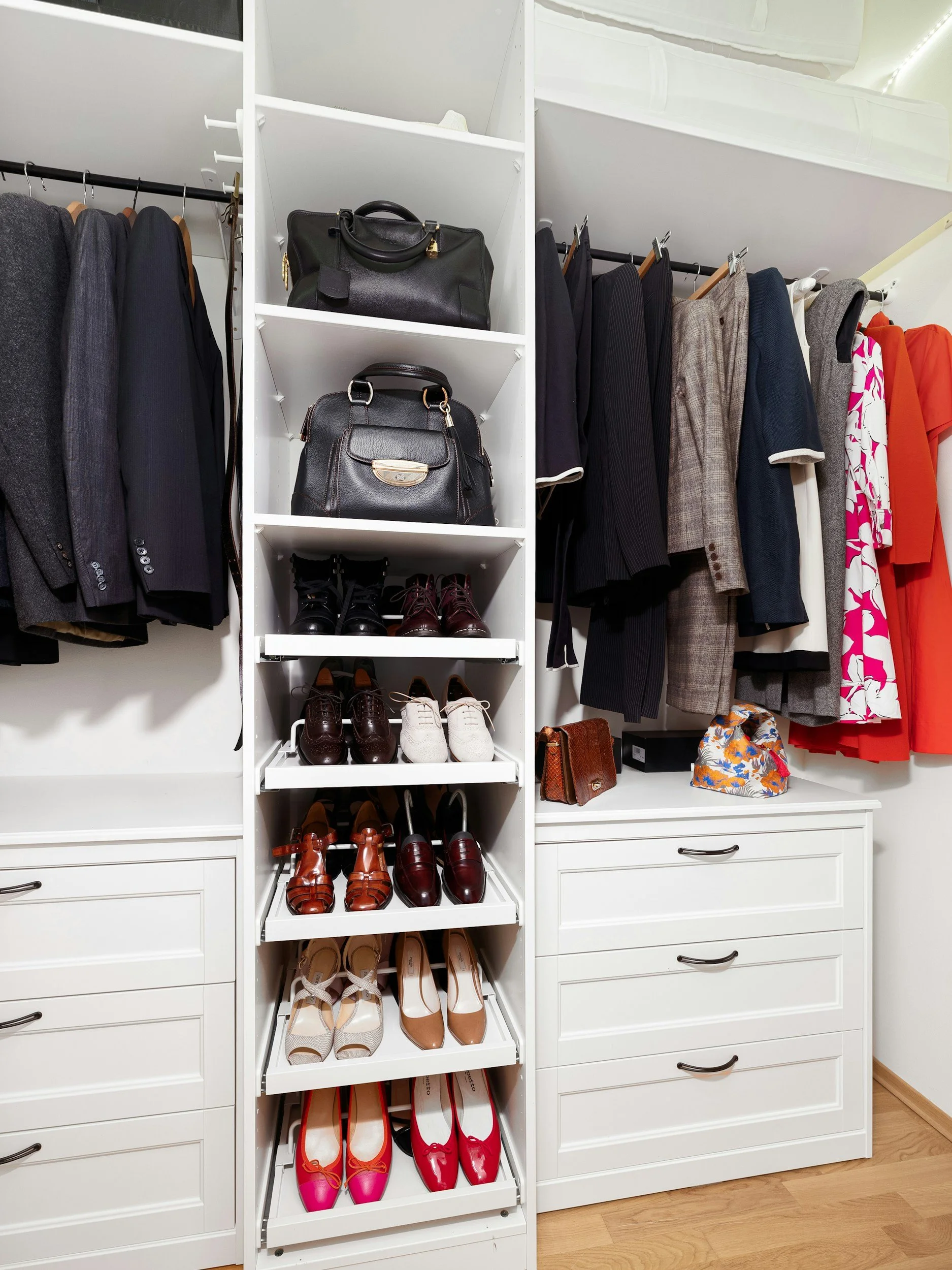 White closet with suits, jackets, and dresses hanging on racks, and shoes, handbags, and a small purse on open shelves and drawers.
