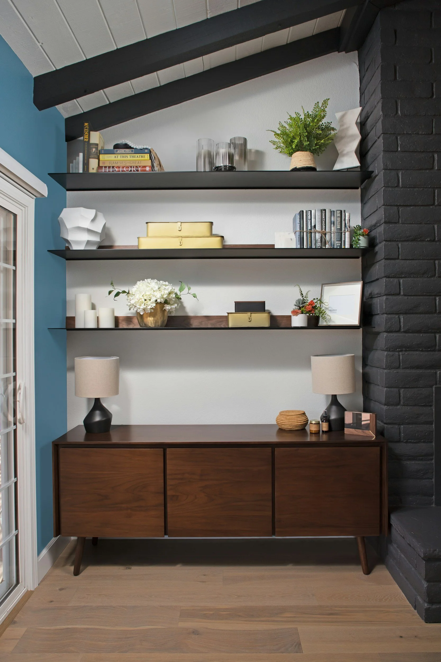 A living room corner with a wooden sideboard, two table lamps, and three black floating shelves with books, decor items, and a potted plant.