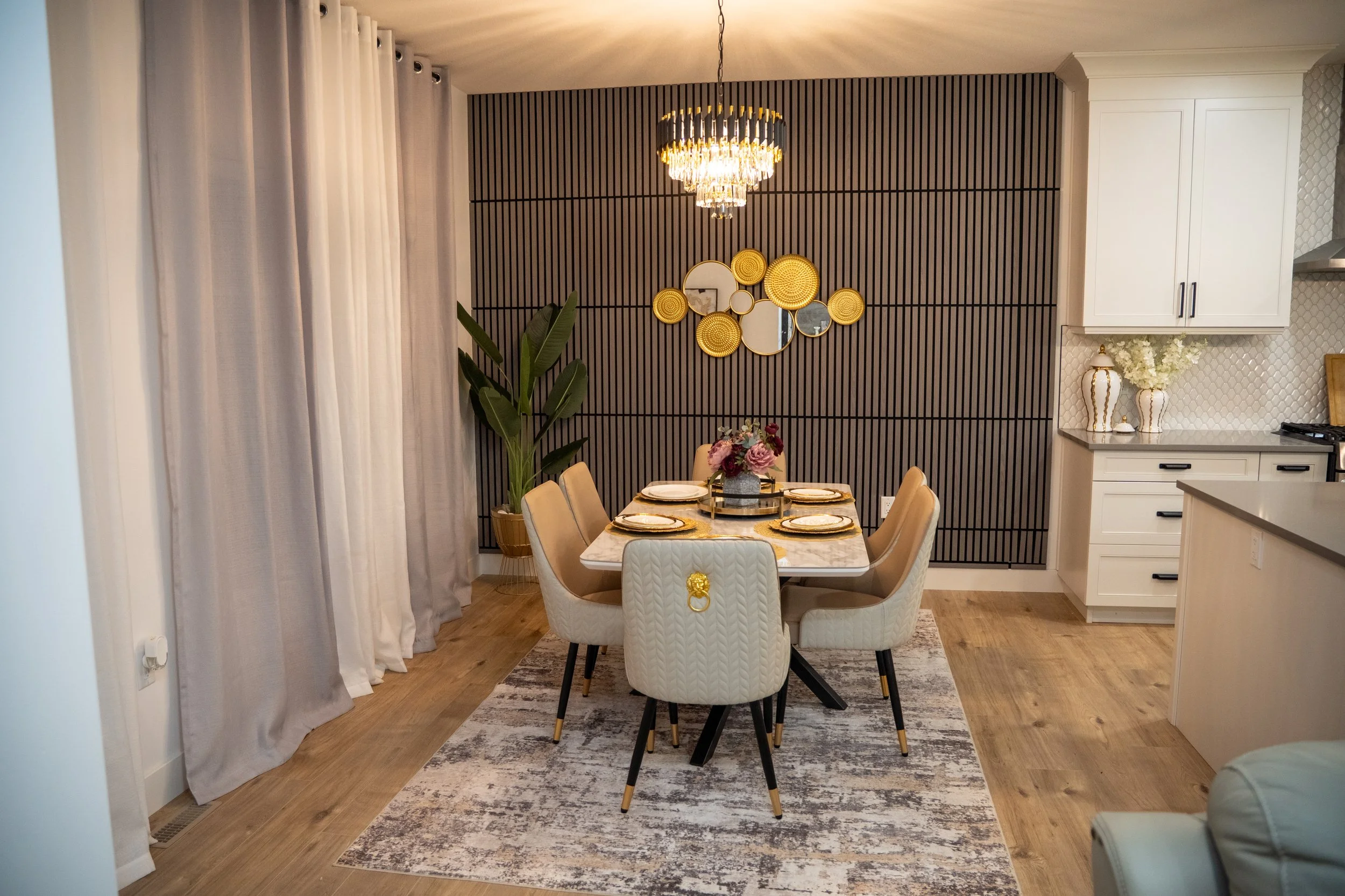 A dining room with a rectangular marble table set with gold-rimmed plates and floral centerpiece, surrounded by six beige upholstered chairs with gold accents, a dark slatted accent wall with round mirrors and woven decor, a modern chandelier overhead, and large curtains on the left side.