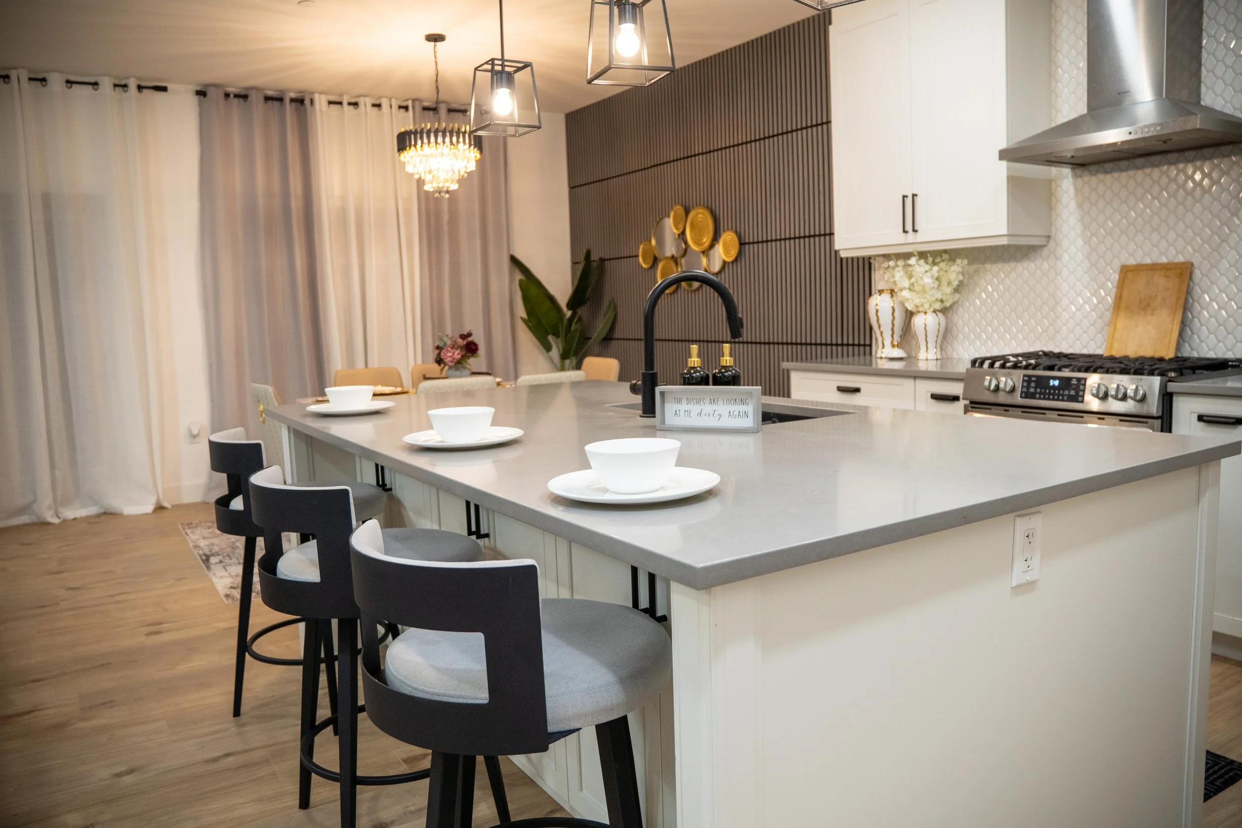 Modern kitchen with a large island countertop set with four white bowls and plates, black and gray bar stools, cabinetry, pendant lights, and decorative wall accents, with a dining area in the background.