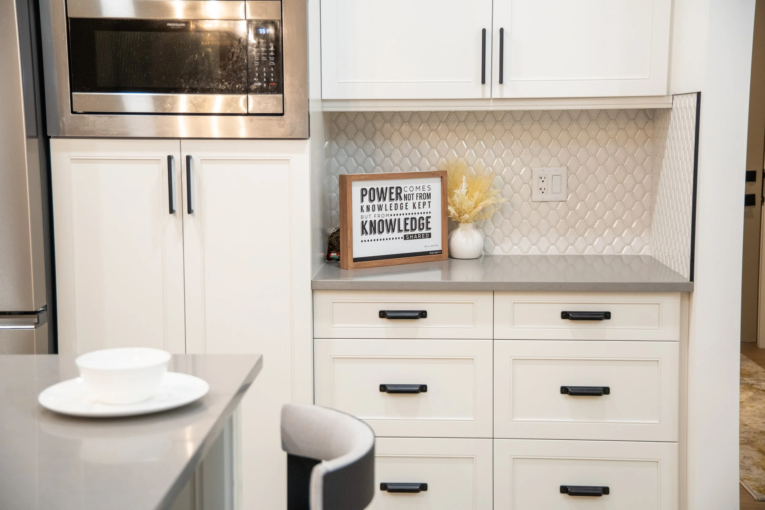 Kitchen with white cabinets, black handles, stainless steel microwave, yellow flower arrangement, framed quote on a shelf, electrical outlet, and a partial view of a table with a bowl and plate.