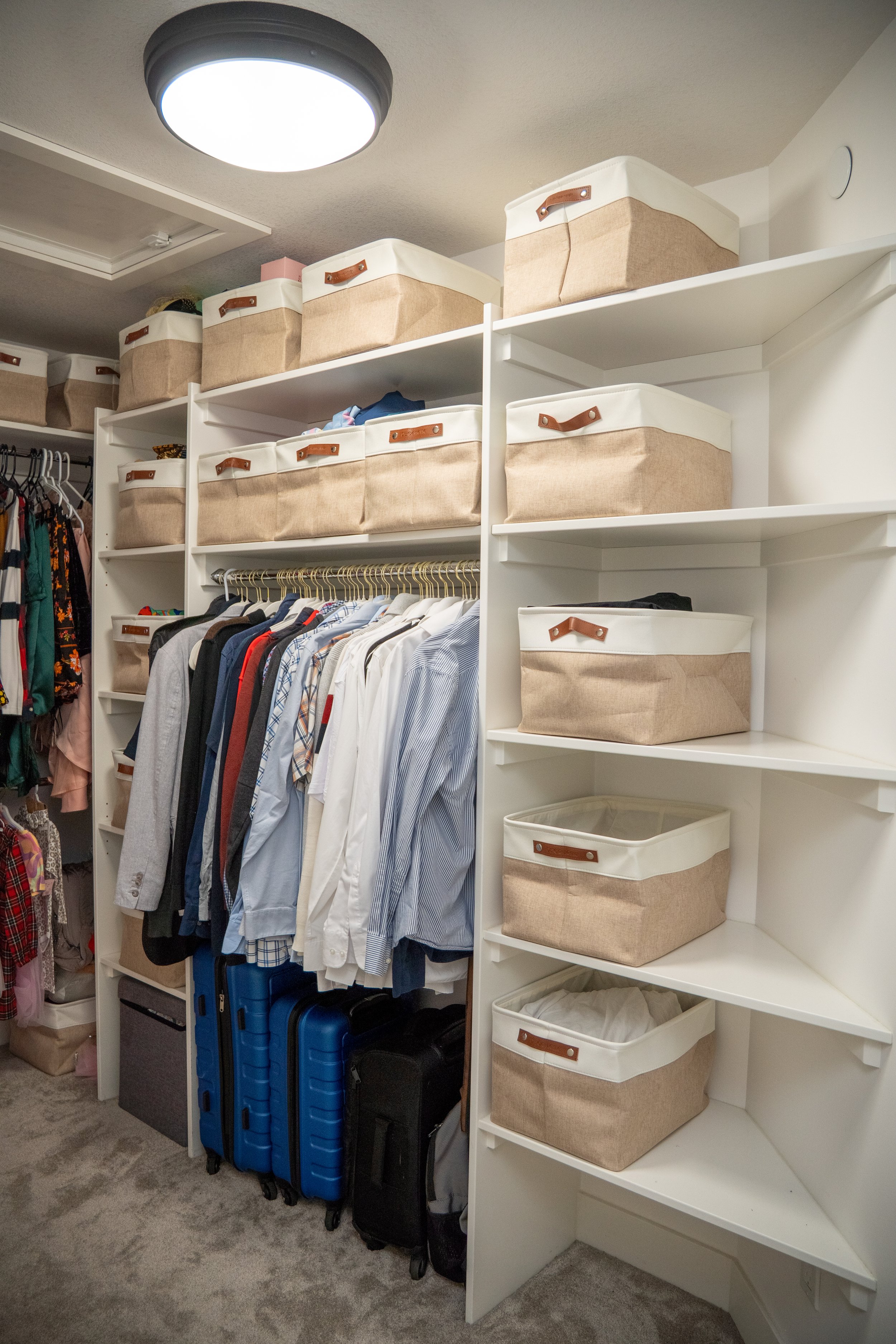Closet with neatly organized white shelving and storage bins, hanging clothes, three blue suitcases, and a clothes rack.