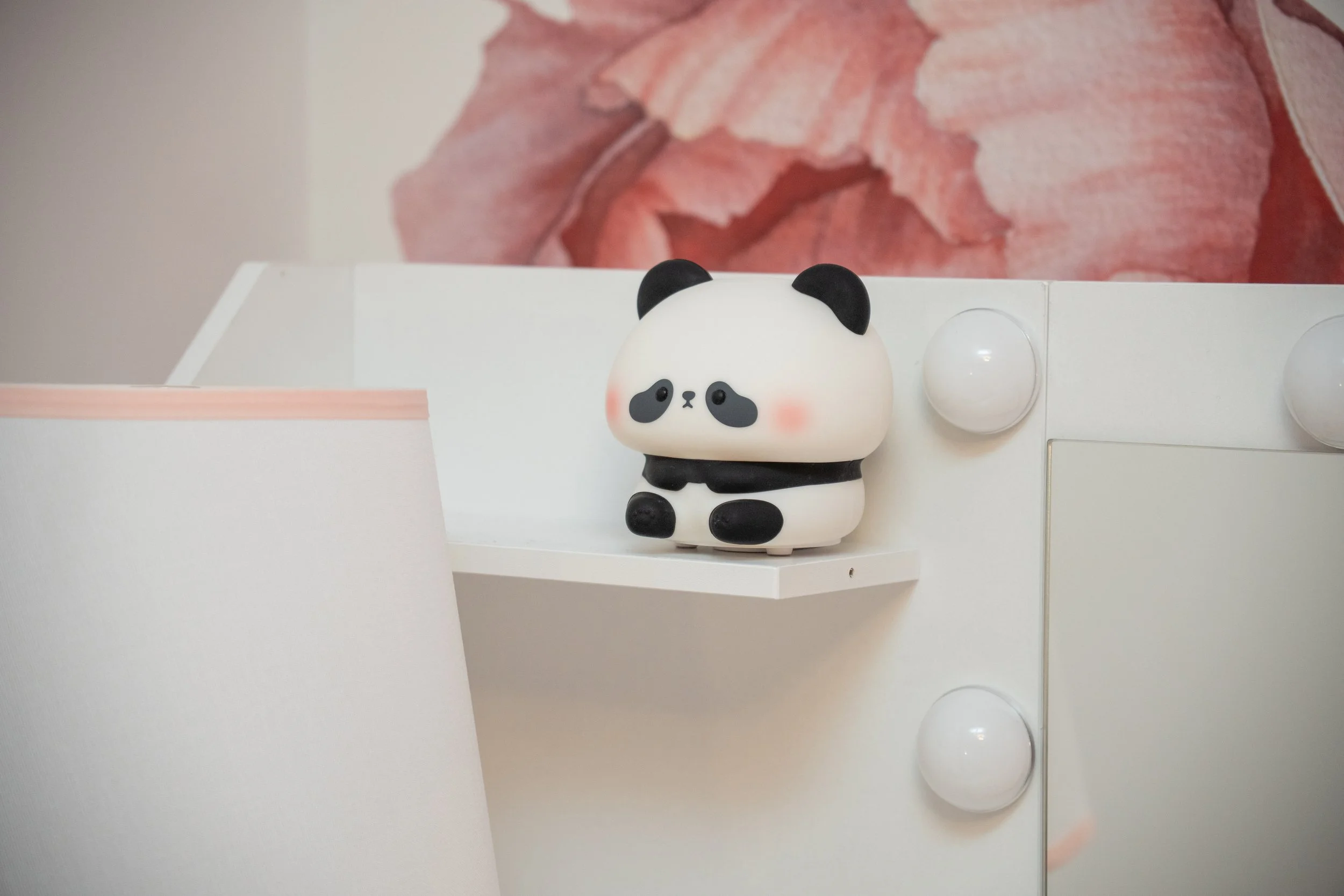 Cute panda-shaped night light with a white body, black ears, and blush cheeks sitting on a white shelf in front of a mirror with light bulbs, with a pink floral background