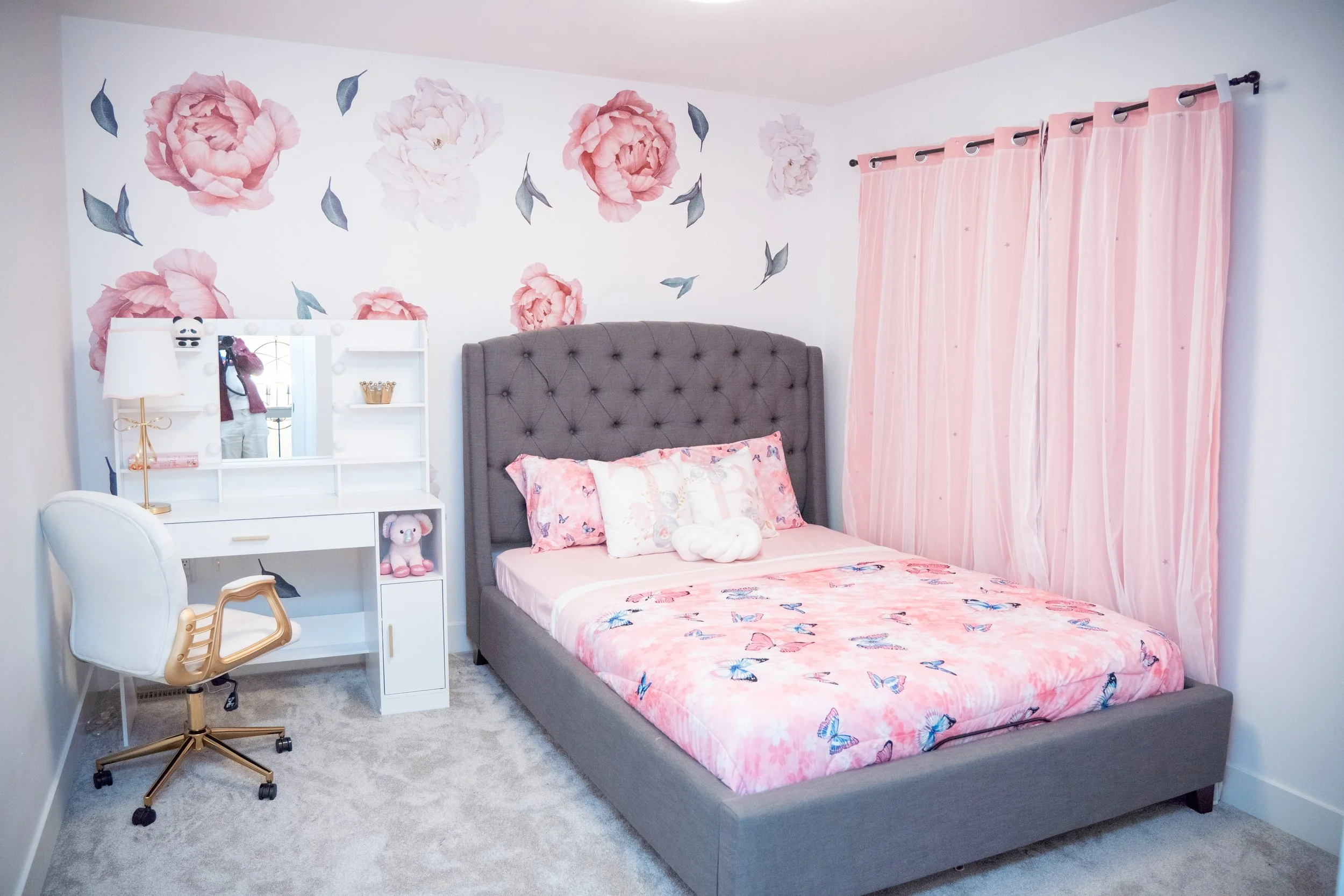 A bedroom with a pink and white floral themed decor, a bed with pink butterfly blanket and stuffed toys, a white desk with mirror and plush toy, and pink curtains.