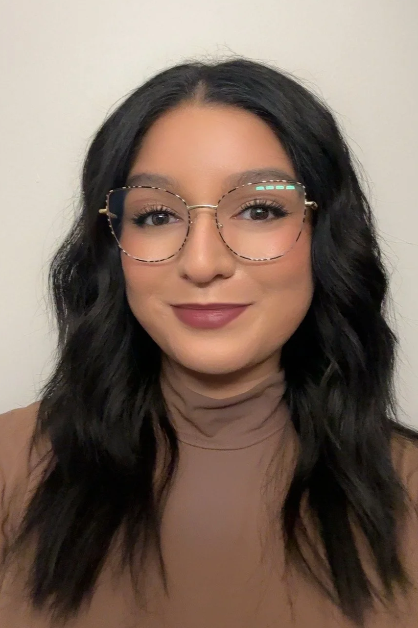 A woman with long black wavy hair wearing glasses and a tan turtleneck
