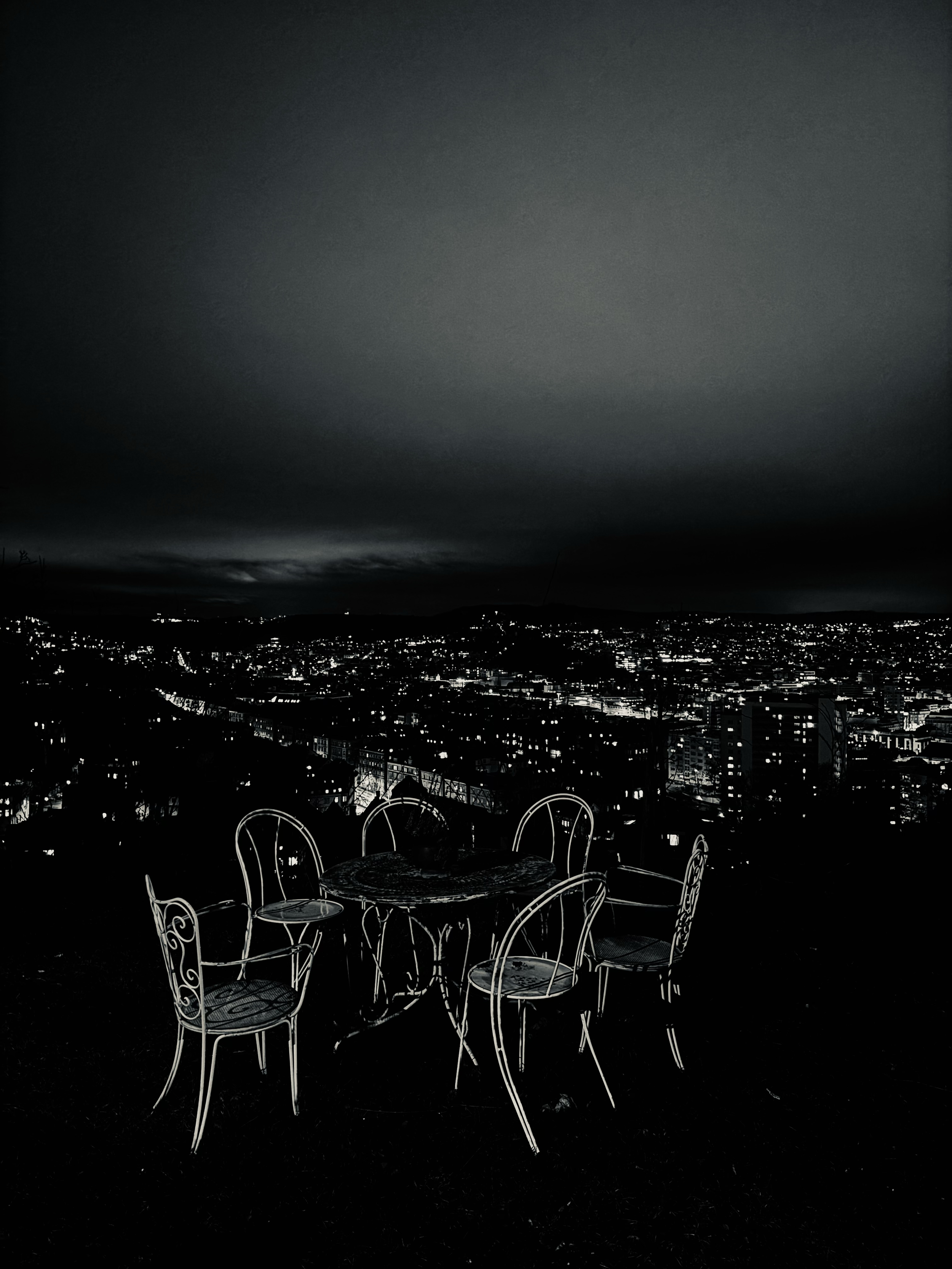 A surreal nighttime scene with empty chairs and a table on a terrace, overlooking the city below, creating the feeling of a quiet ghostly dinner. Eine surreale Nachtszene mit leeren Stühlen und einem Tisch auf einer Terrasse mit Blick über die Stadt,