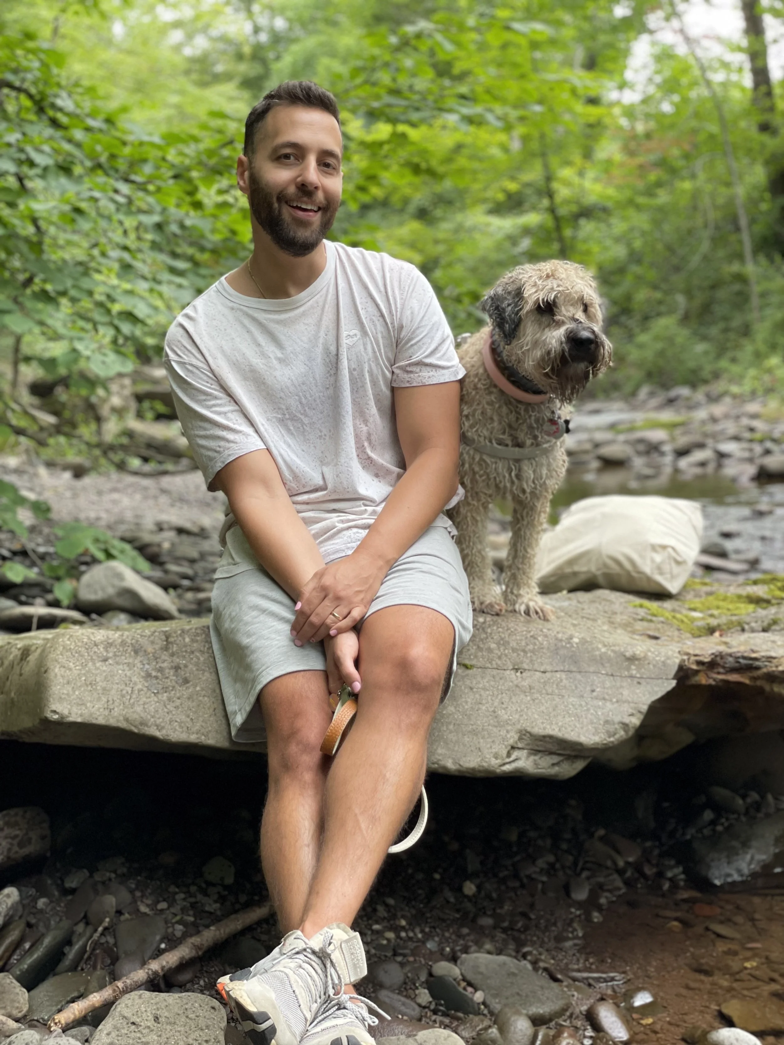 A smiling man sitting on a large rock in a forest, holding a leash attached to a large, fluffy, light-colored dog with a pink collar, beside a stream surrounded by green foliage.