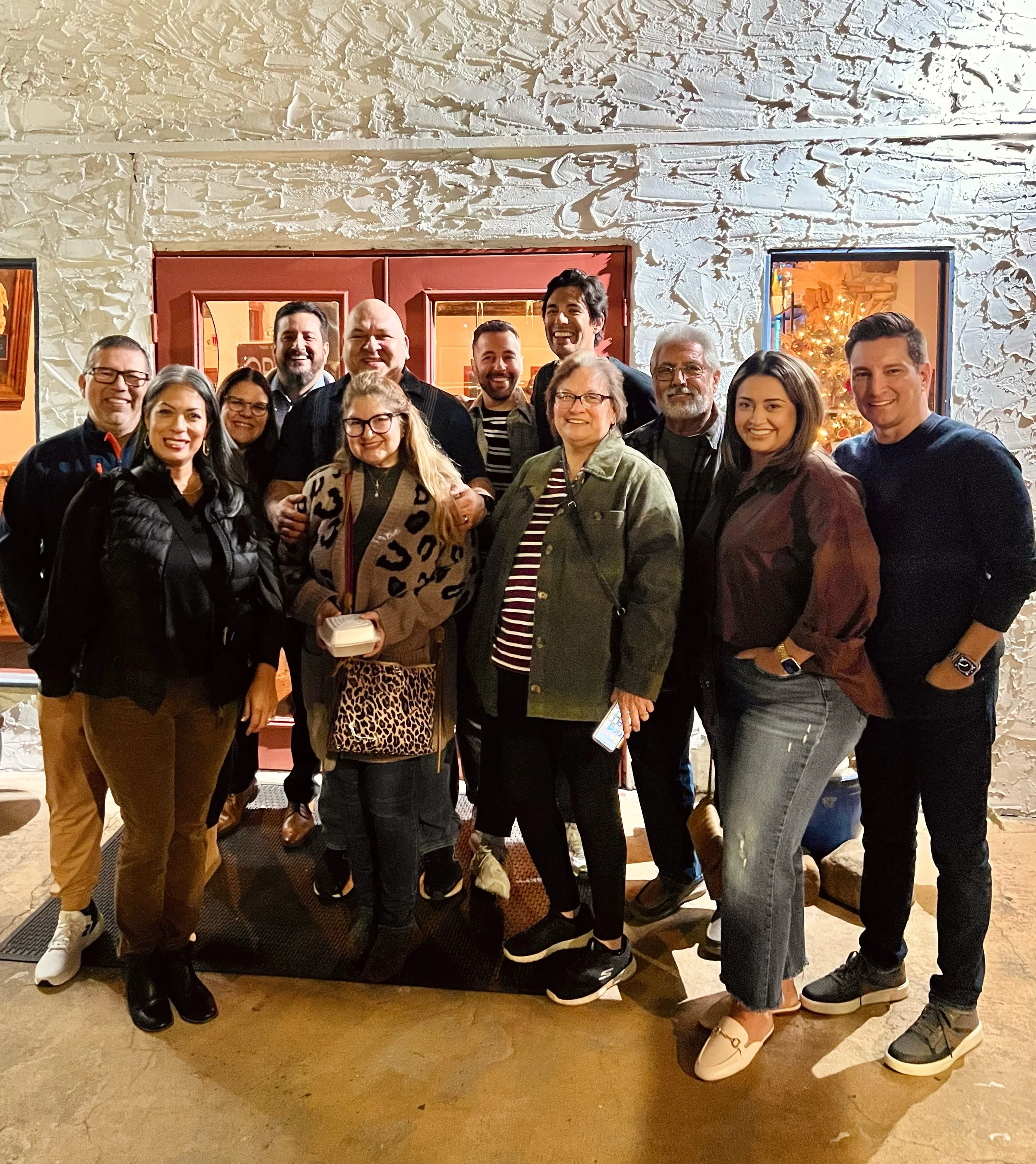 A group of twelve people smiling, standing together in front of a red door, with a decorated Christmas tree visible through a window on the right.