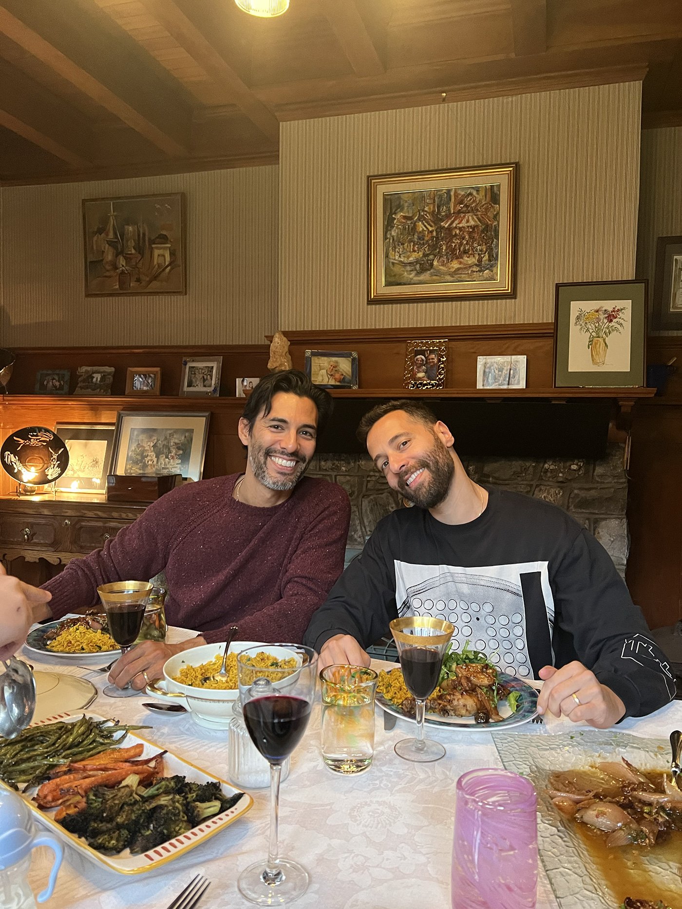 Two men smiling and sitting at a dinner table with plates of food, glasses of wine, and various dishes in a cozy, decorated room.