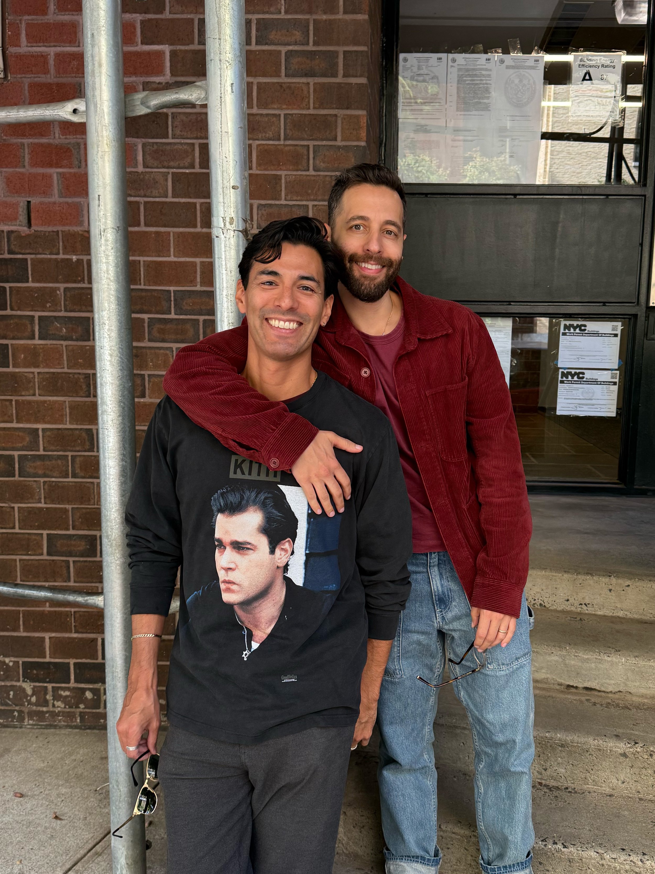 Two men standing close together outside in front of a brick wall and window, smiling for the photo. One is wearing a black shirt with a large image of a man with slicked-back hair, while the other is dressed in a red jacket and jeans.