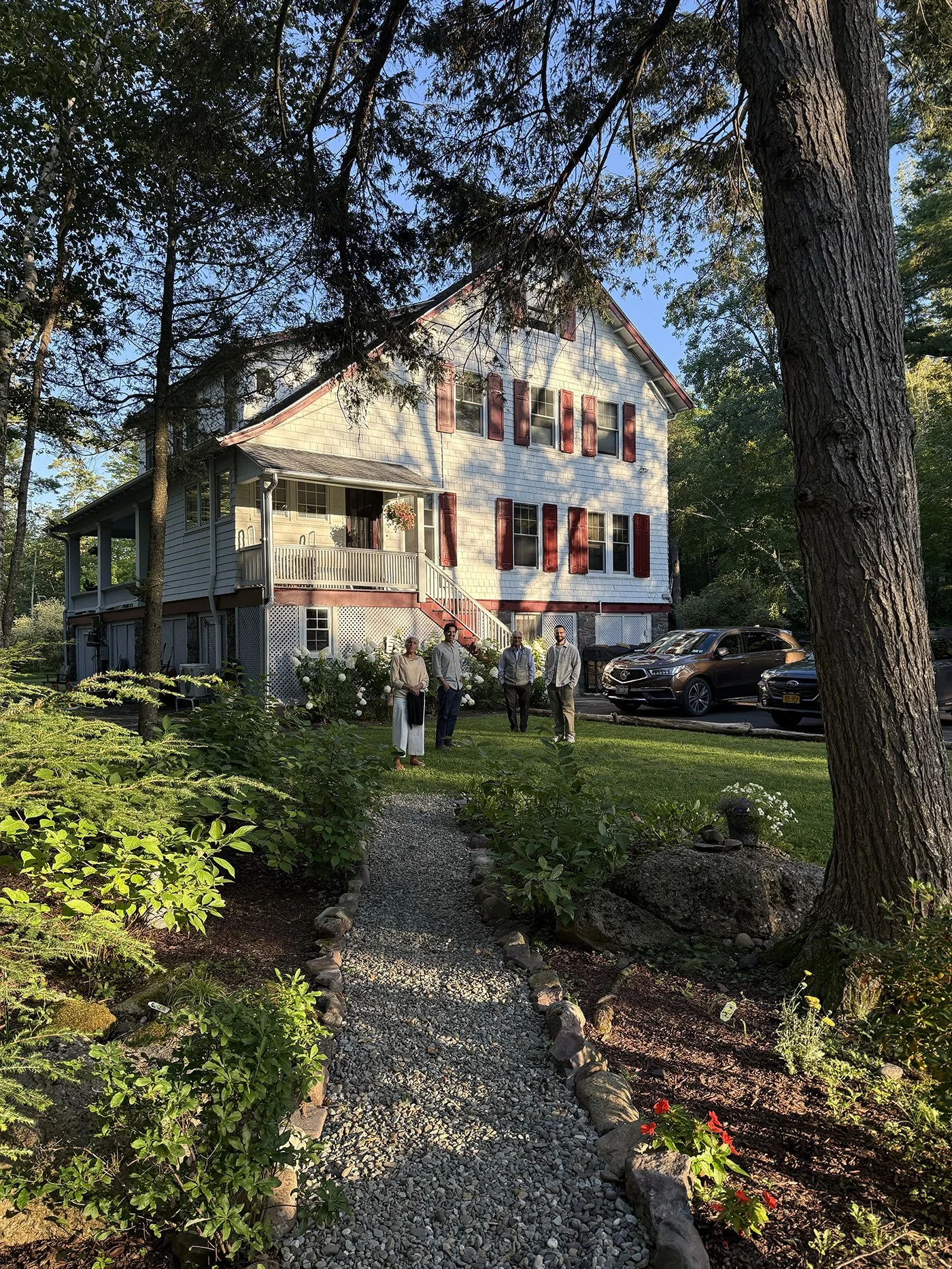 A group of five people standing on a lawn in front of a large, white, multi-story house with red shutters and a porch. There are trees and bushes surrounding the yard, and a gravel path leads to the house. Several parked cars are visible to the right