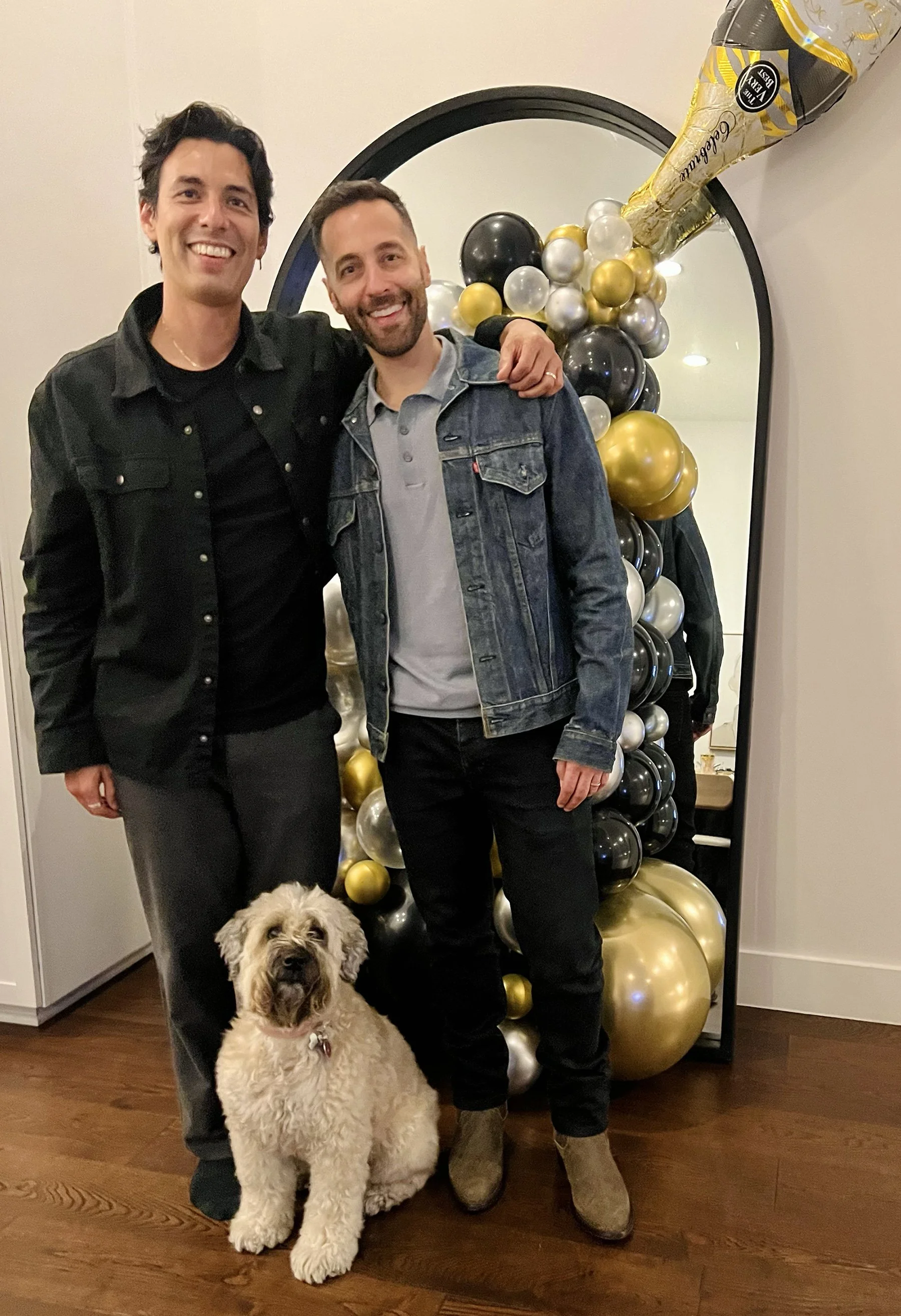 Two men smiling and posing together, one with his arm around the other, standing in front of a decorative balloon arrangement with black, gold, and silver balloons and a large champagne bottle prop. A dog sits in front of them on the wooden floor.