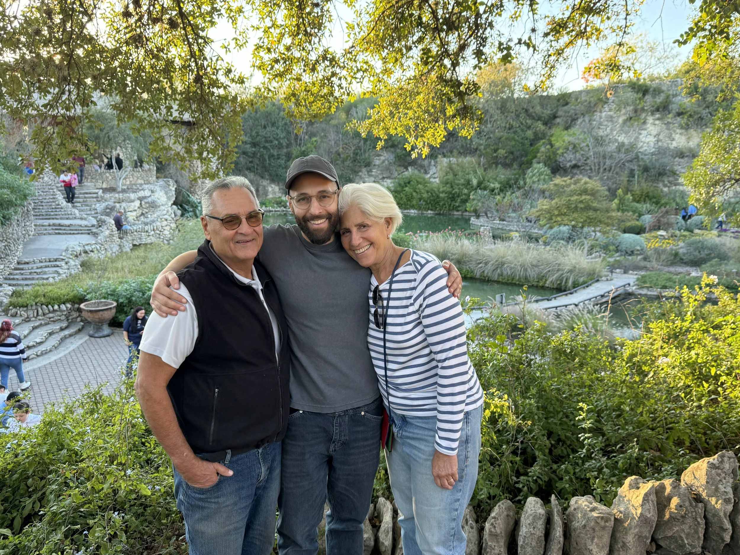 Three smiling people, two men and one woman, standing close together in a park-like setting with greenery, water features, and stone stairs in the background.