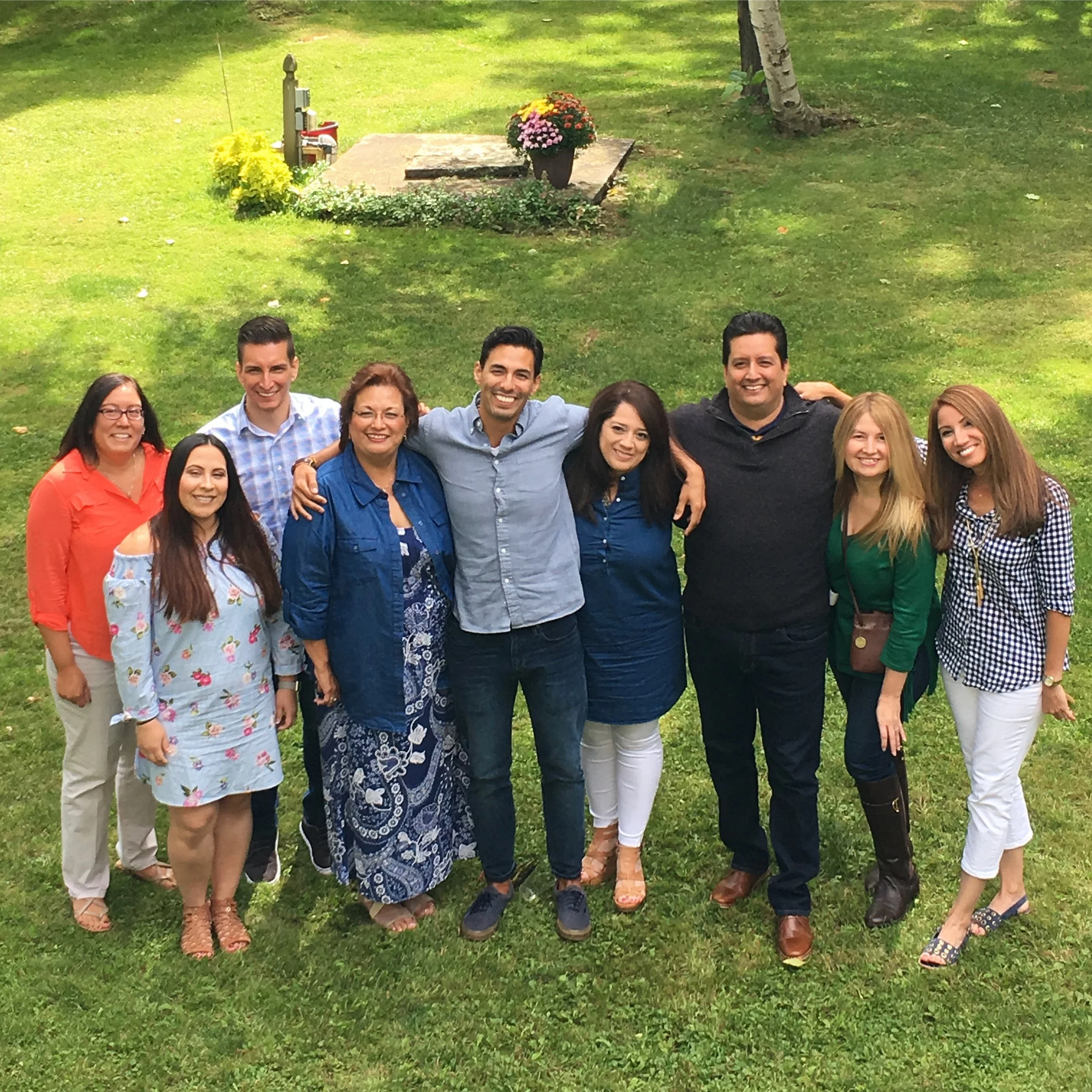 Group of ten people standing outdoors on grass, smiling, with trees and a flower bed in the background.