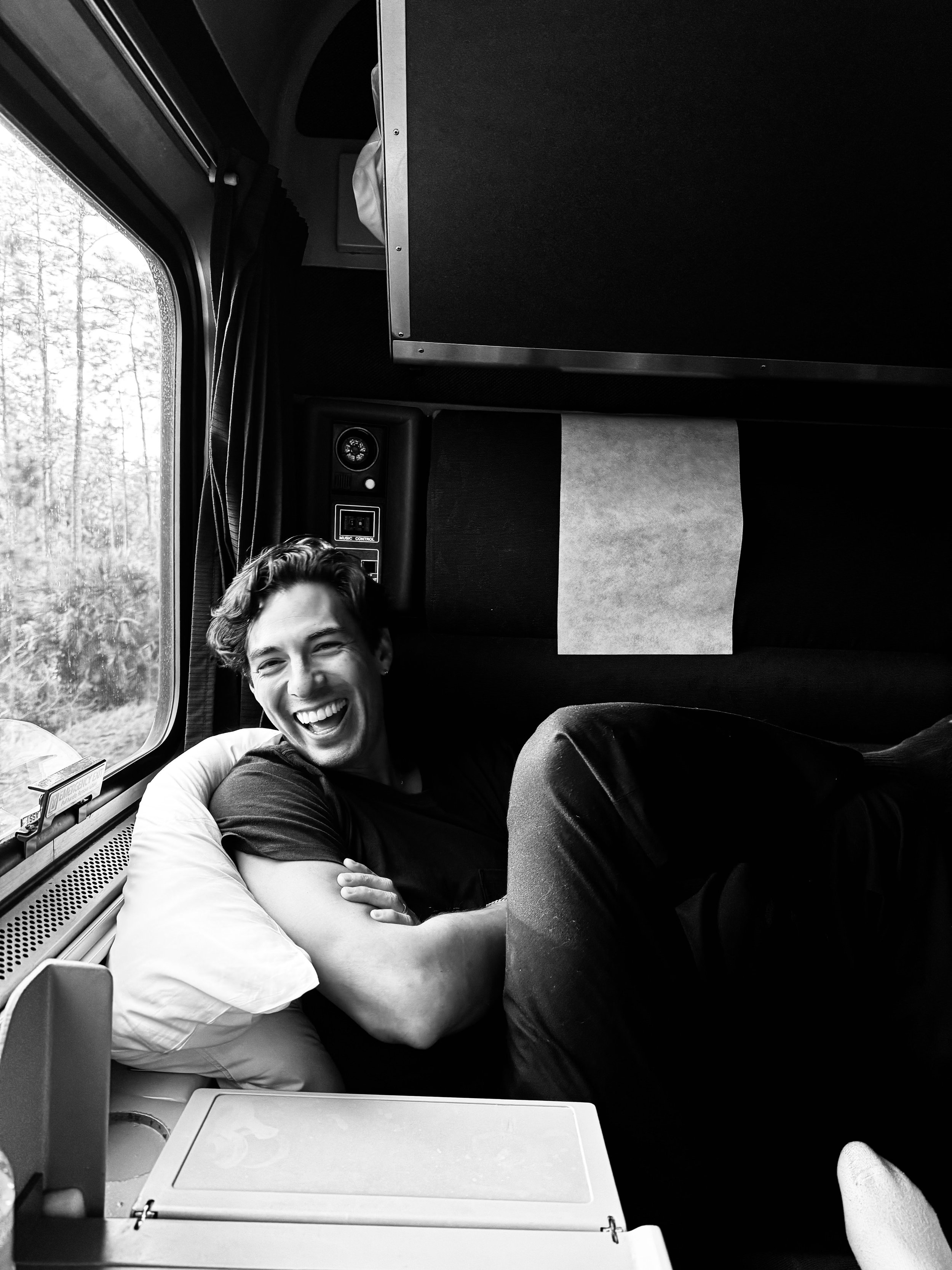 A young man with a broad smile, lying on a pillow, relaxed inside a train or bus with a window showing trees outside.