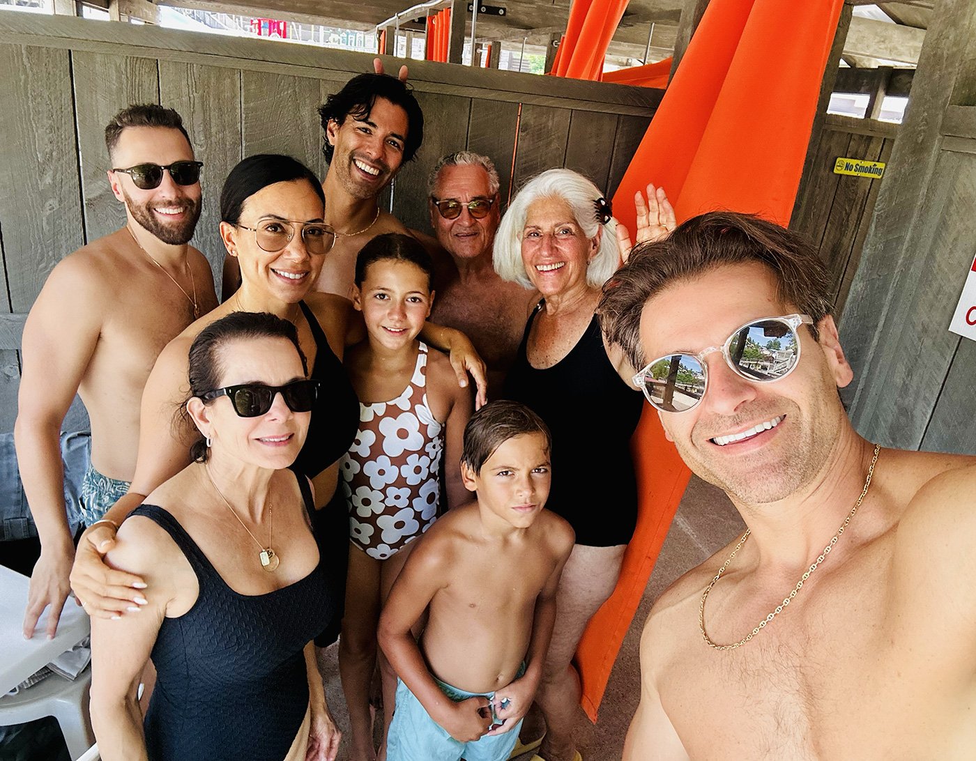 Group of people in swimsuits and summer clothes posing for a selfie at a poolside or beach setting. They are smiling, some wearing sunglasses, with a wooden fence and orange shade structure in the background.