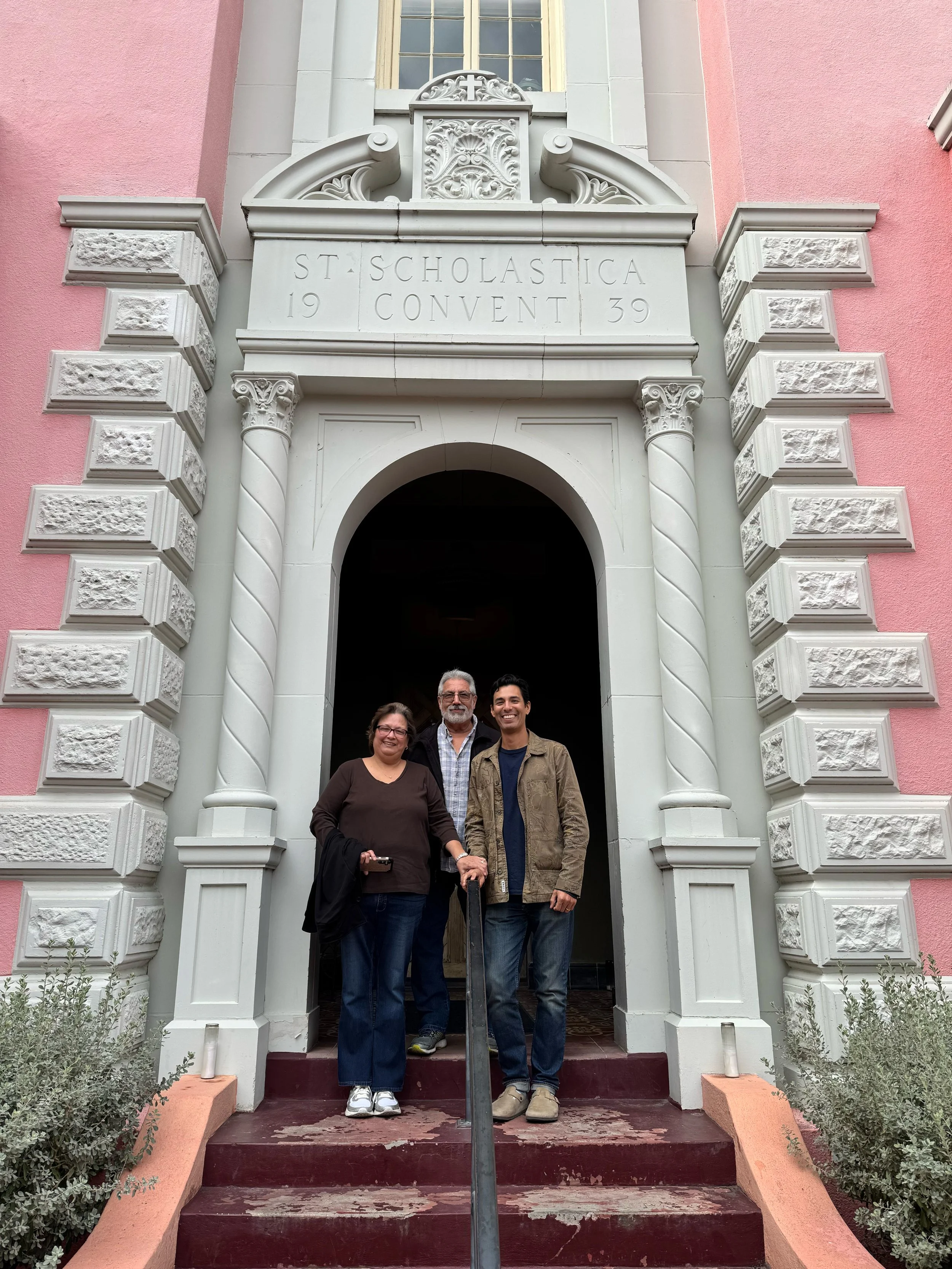 Three people standing on steps in front of the entrance to a historic building with ornate white trim and pink walls, featuring a sign that reads 'St. Scholastica 1939 Convent.'