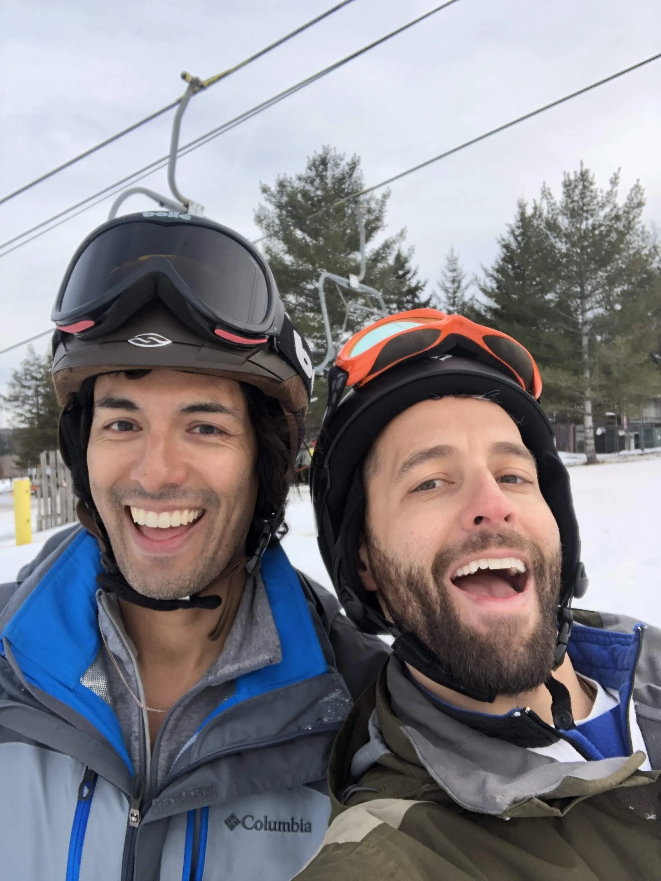 Two men smiling outdoors in a snowy area, wearing helmets and winter jackets, with ski lift cables and trees in the background.