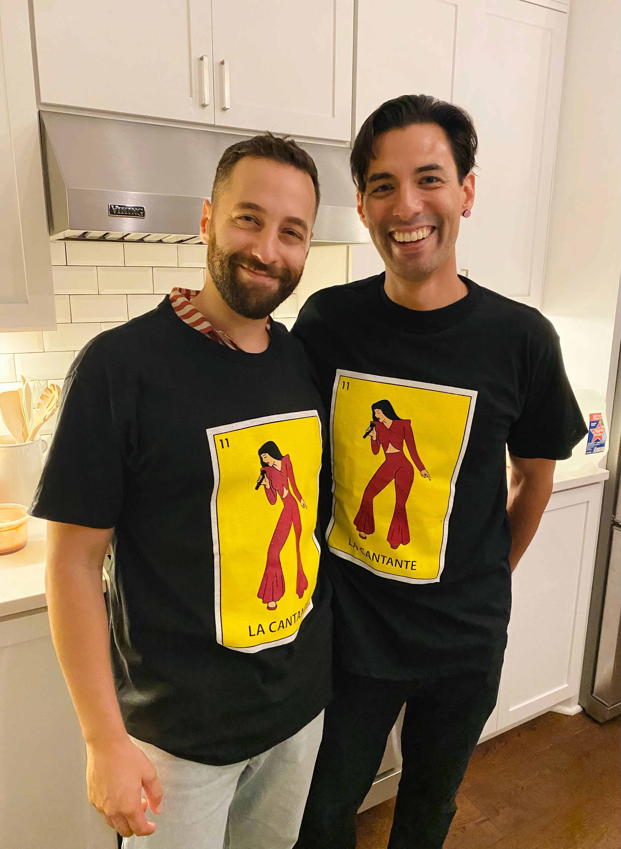 Two men standing in a kitchen smiling and wearing matching black t-shirts with a yellow design of a singer holding a microphone, labeled 'La Cantante.'