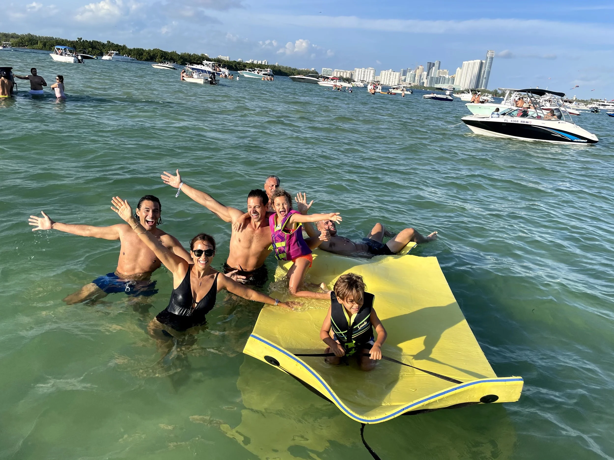 Group of smiling people enjoying swimming and relaxing on a yellow floating mat in a waterfront area with boats and city skyline in the background.