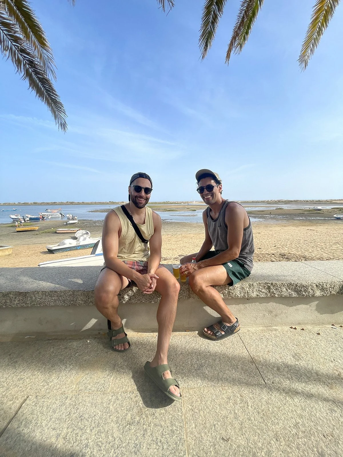 Two men sitting on a stone ledge at the beach, smiling, with small boats anchored in the water behind them, palm trees overhead, clear blue sky.