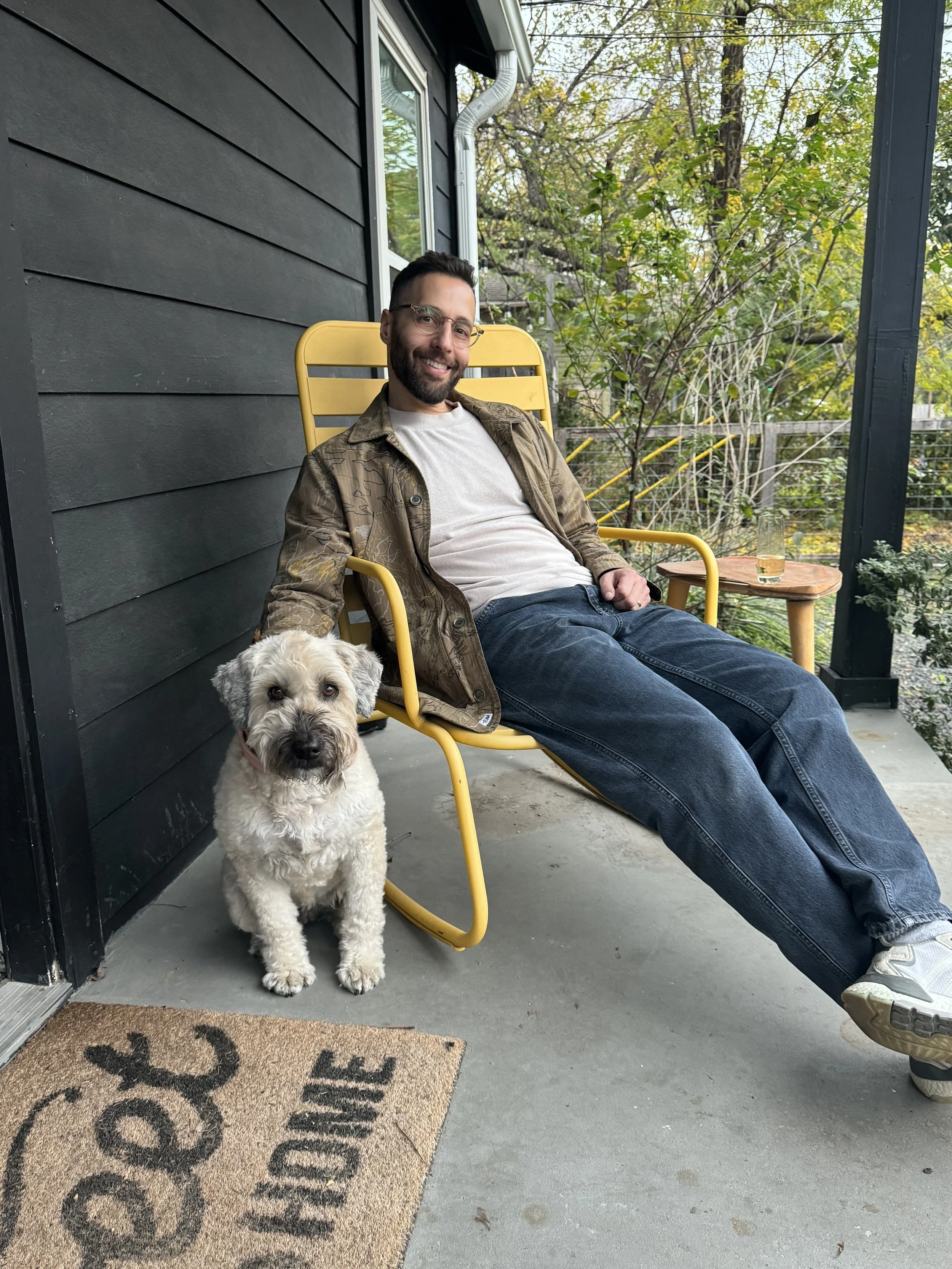 A man with glasses and a beard sits on a yellow outdoor chair on a porch next to a white and gray dog. There is a small wooden table with a glass of water on it, and greenery in the background.
