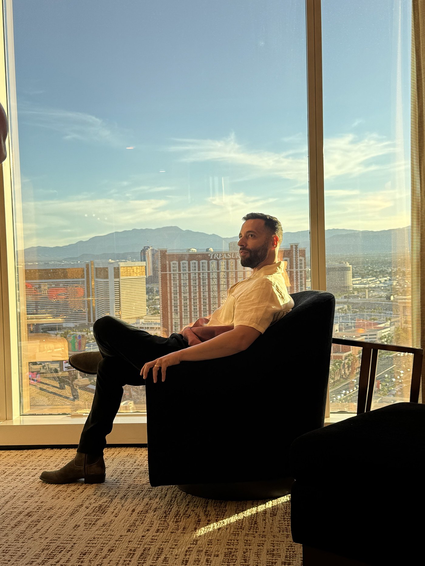 A man sitting in a black chair by a large window with a city view and mountains in the background, during sunset or sunrise.