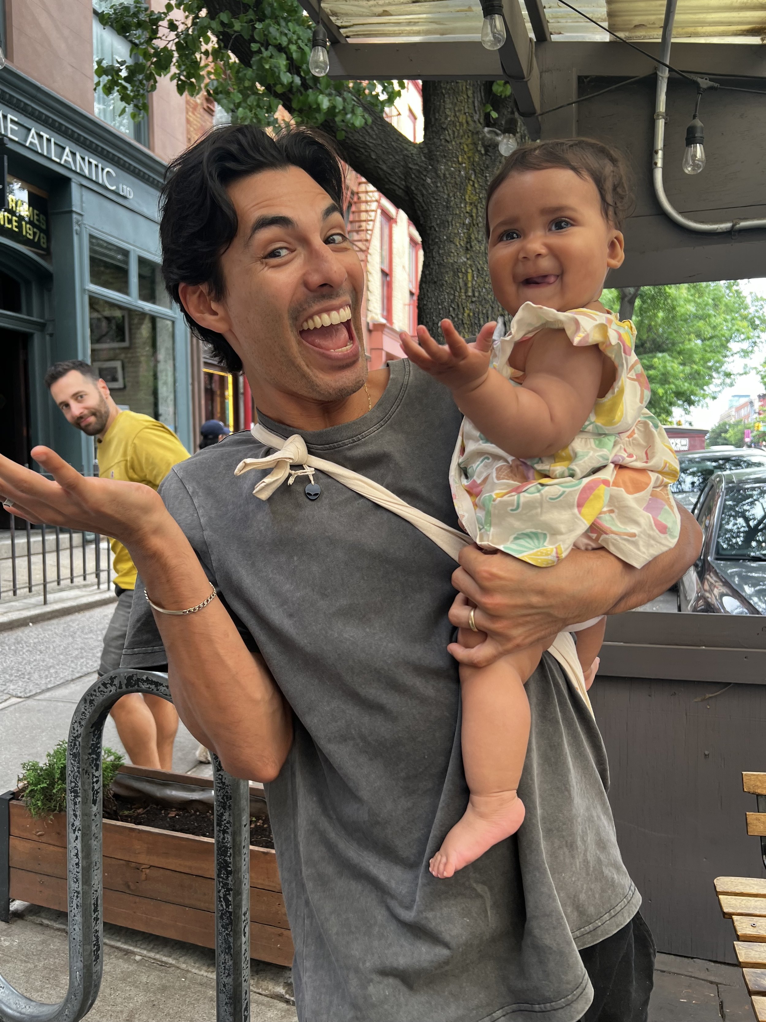 A man smiling and showing a playful gesture while holding a happy baby girl on his shoulder. The baby is smiling and waving, wearing a colorful dress, outdoors on a city street.