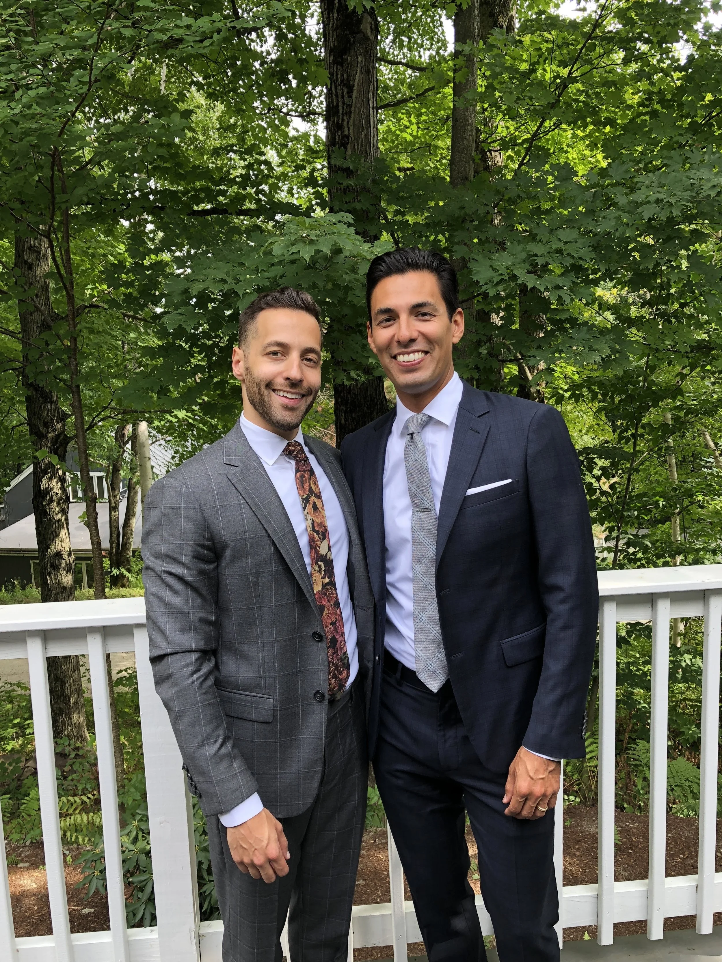 Two men in suits smiling and standing side by side outdoors on a balcony with a white railing, surrounded by green trees.