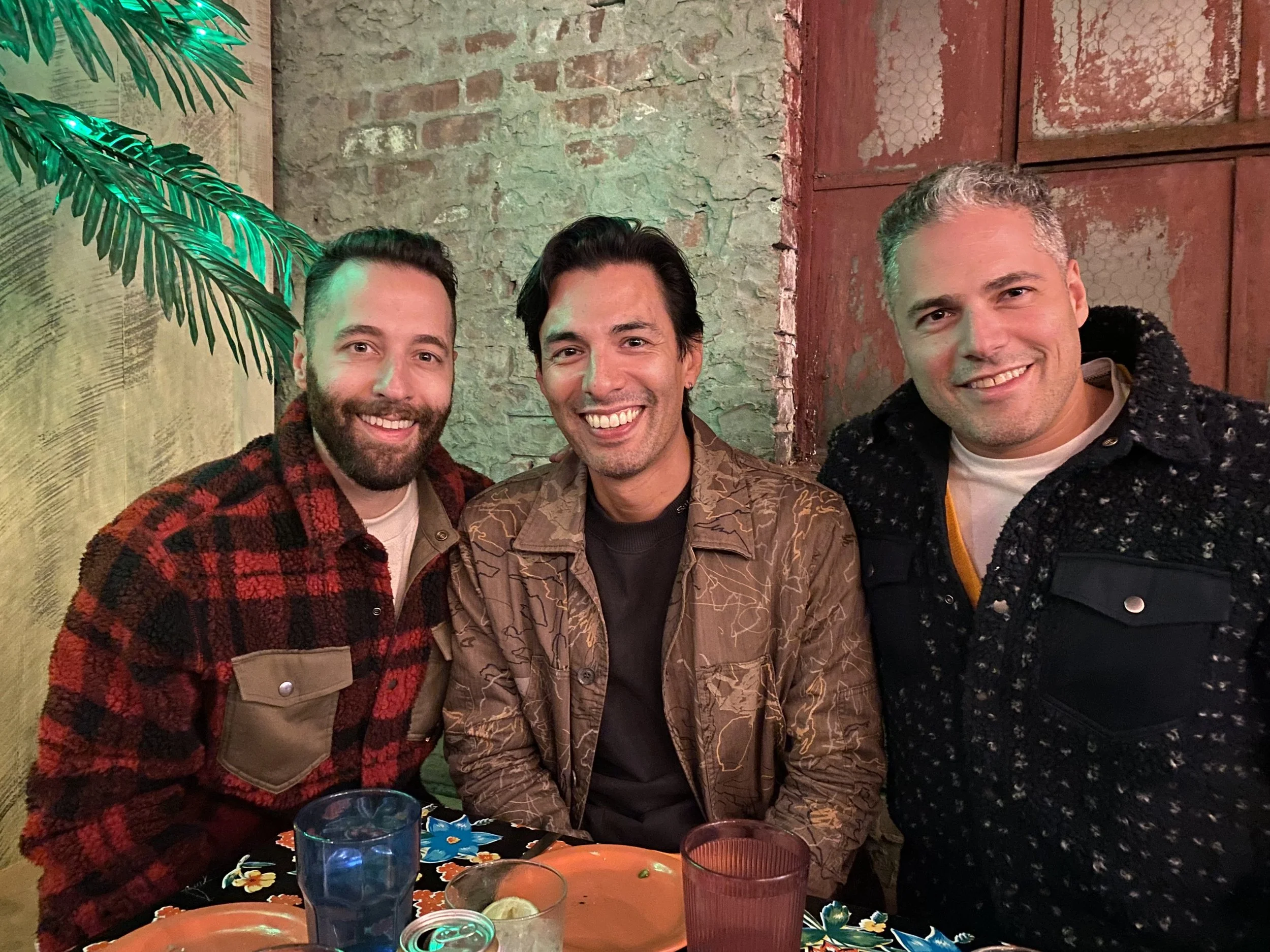 Three men smiling at a table in a cozy, rustic restaurant with a brick wall and a large green plant in the background.