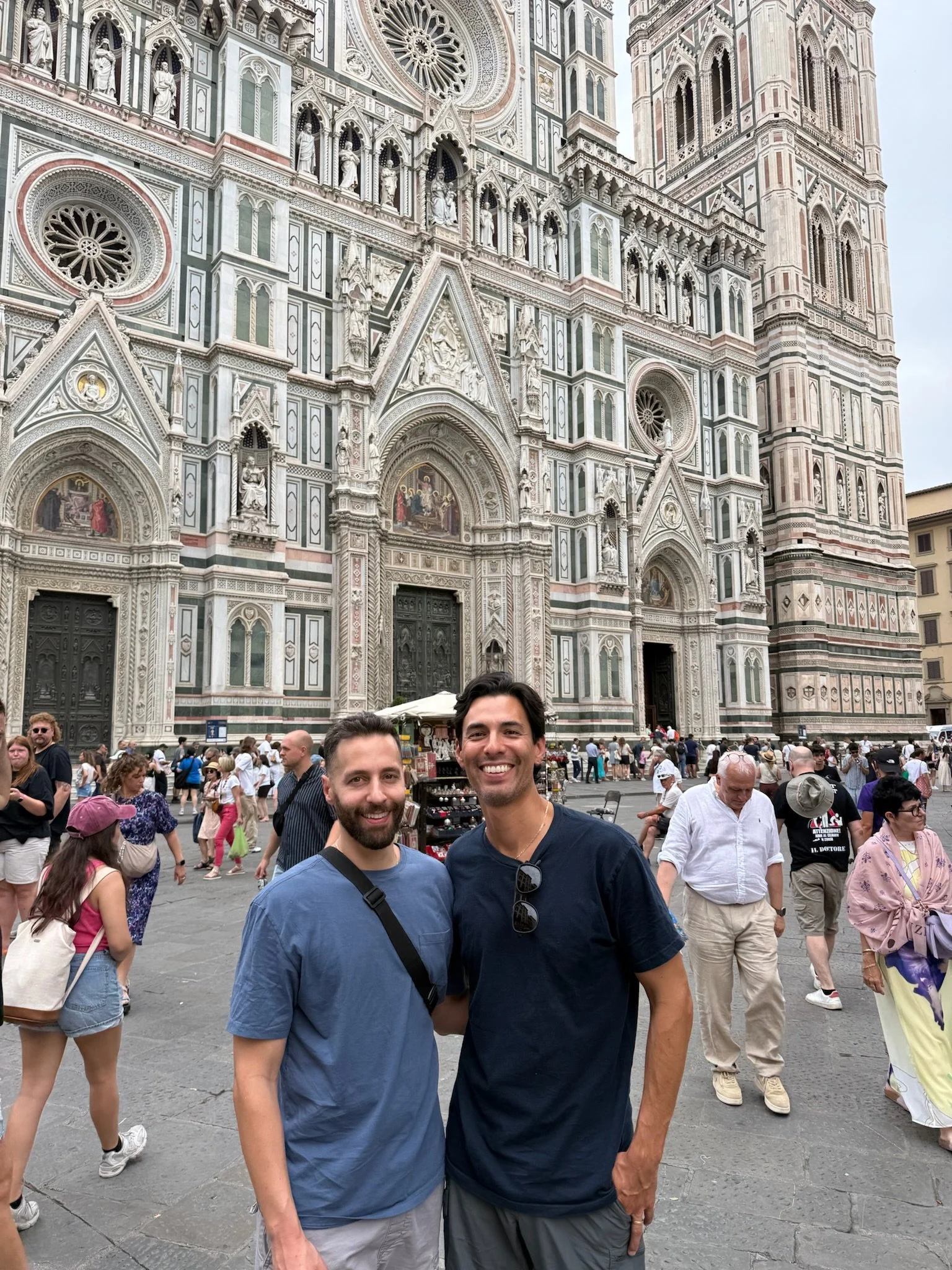 Two men smiling in front of Florence Cathedral in Italy with the cathedral's intricate white, green, and pink marble facade in the background, and other tourists around.
