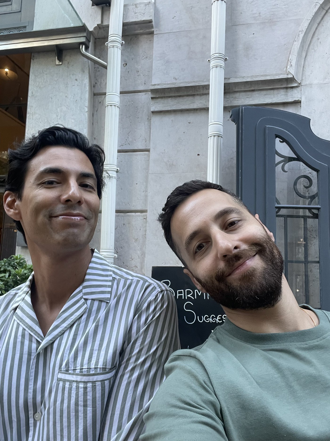 Two men taking a selfie outdoors in front of a building with a sign that reads 'SUGGESTIONS'.