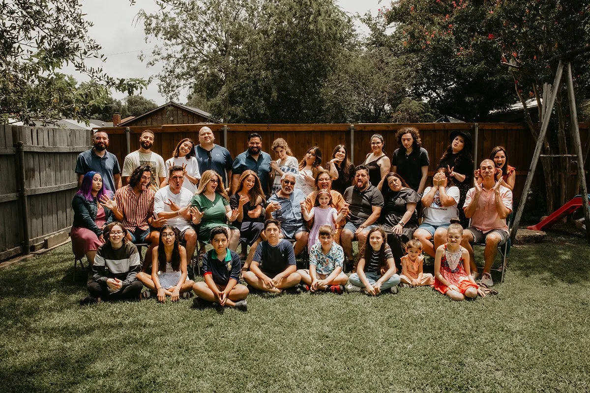 A large group of people, including children and adults, posed for a photo in a backyard with a wooden fence and trees in the background. Some people are sitting on chairs, while others are standing behind them on the grass.