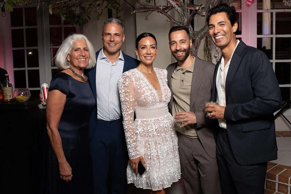 Group of five people smiling and posing for a photo at a social gathering indoors, with a tree and windows in the background.