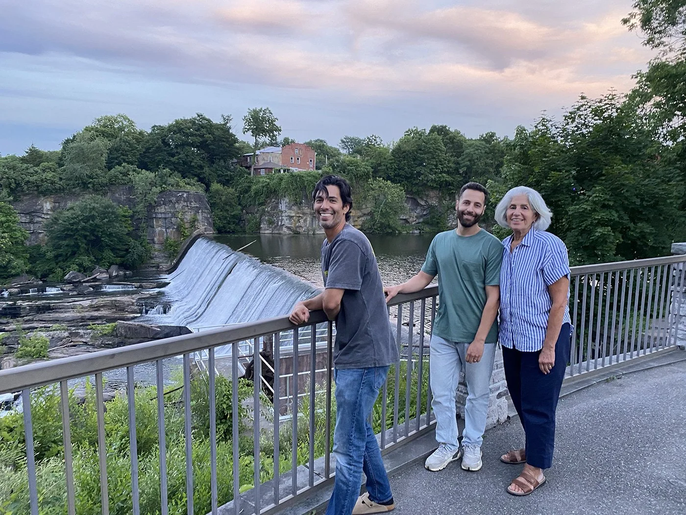 Three people smiling on a bridge overlooking a river with a small waterfall, surrounded by green trees and houses in the background at sunset.