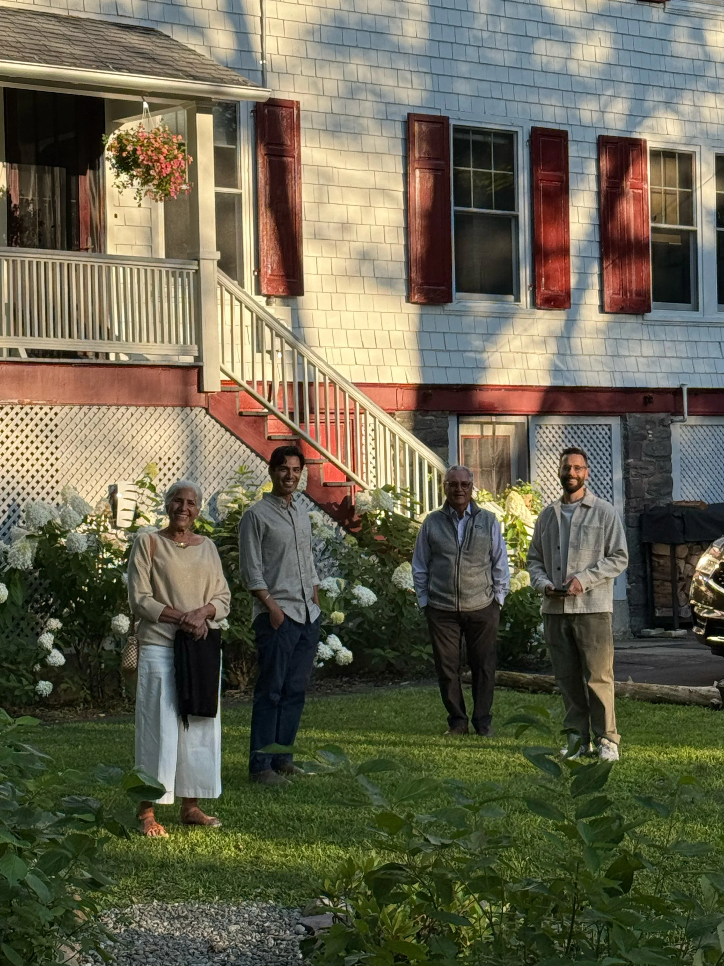 Four people standing on a lawn in front of a house with a staircase, garden plants, and windows with red shutters.