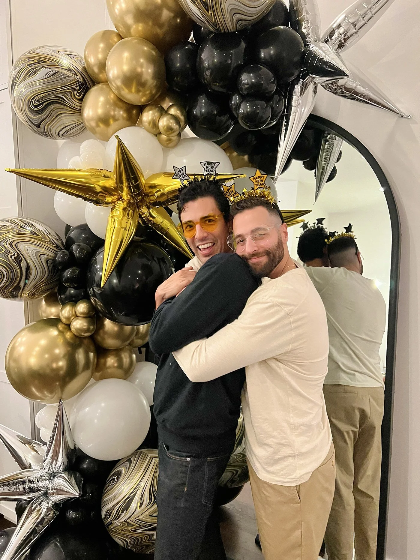 Two men hugging in front of a balloon decorations featuring black, gold, white, and marbled balloons with star-shaped balloons reading 'Happy New Year' and 'Happy' for a celebration.