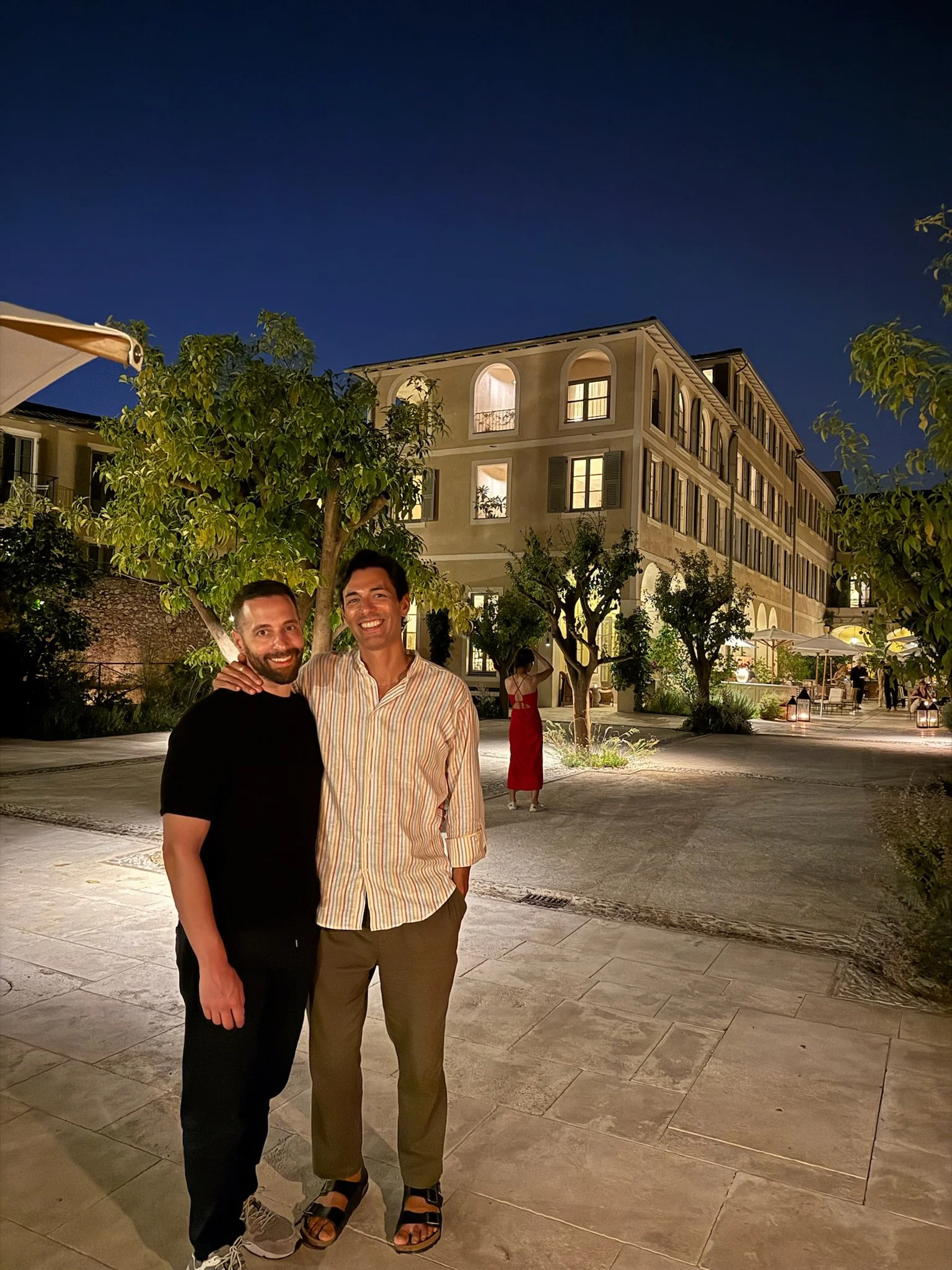 Two men standing together at night in an outdoor courtyard with a multi-story building in the background, trees, and warm lighting.