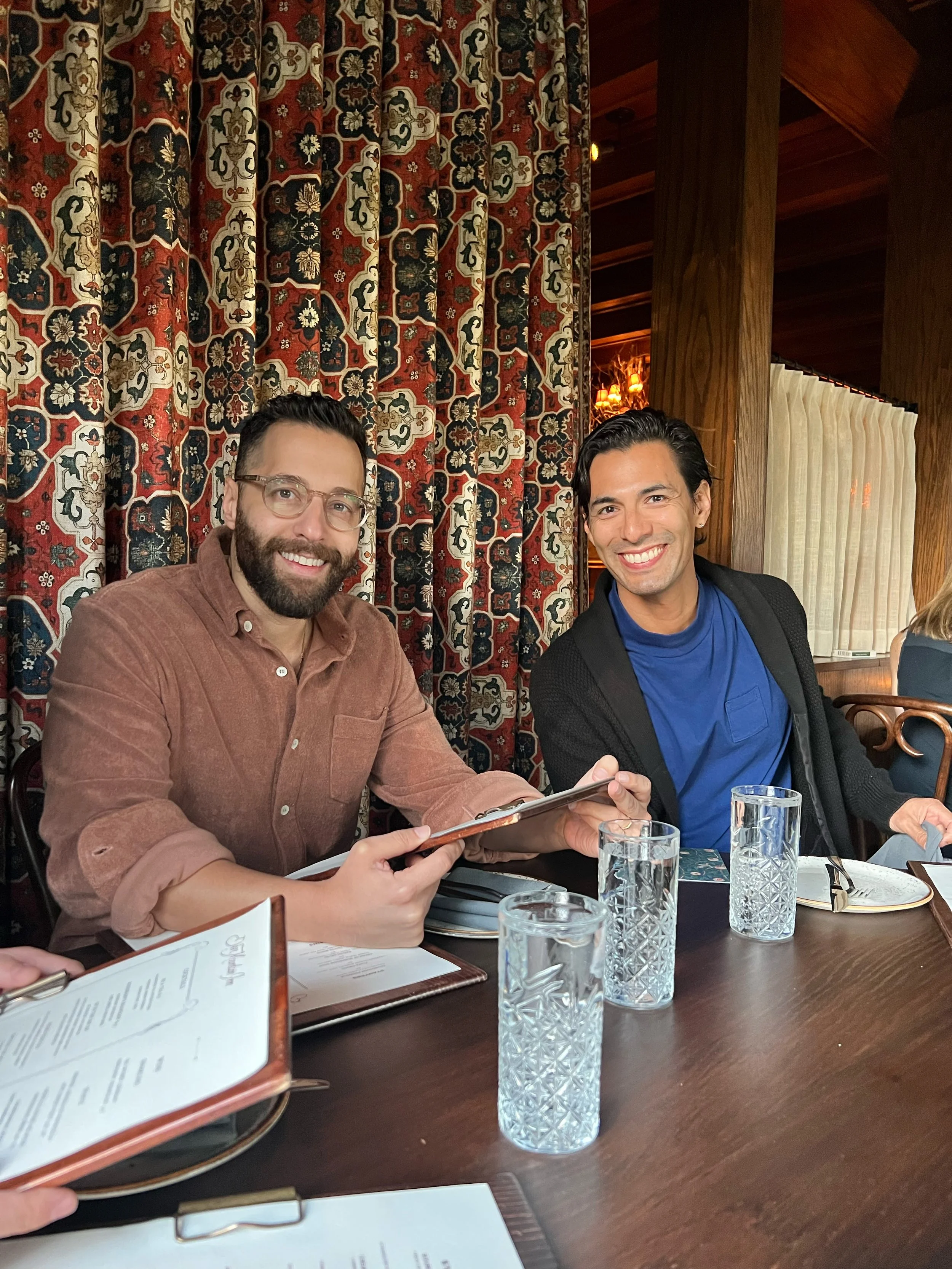 Two men sitting at a restaurant table, smiling, with menus and glasses of water in front of them. The man on the left has glasses and a beard, wearing a brown shirt, and the man on the right has dark hair and a bright smile, wearing a blue shirt and 
