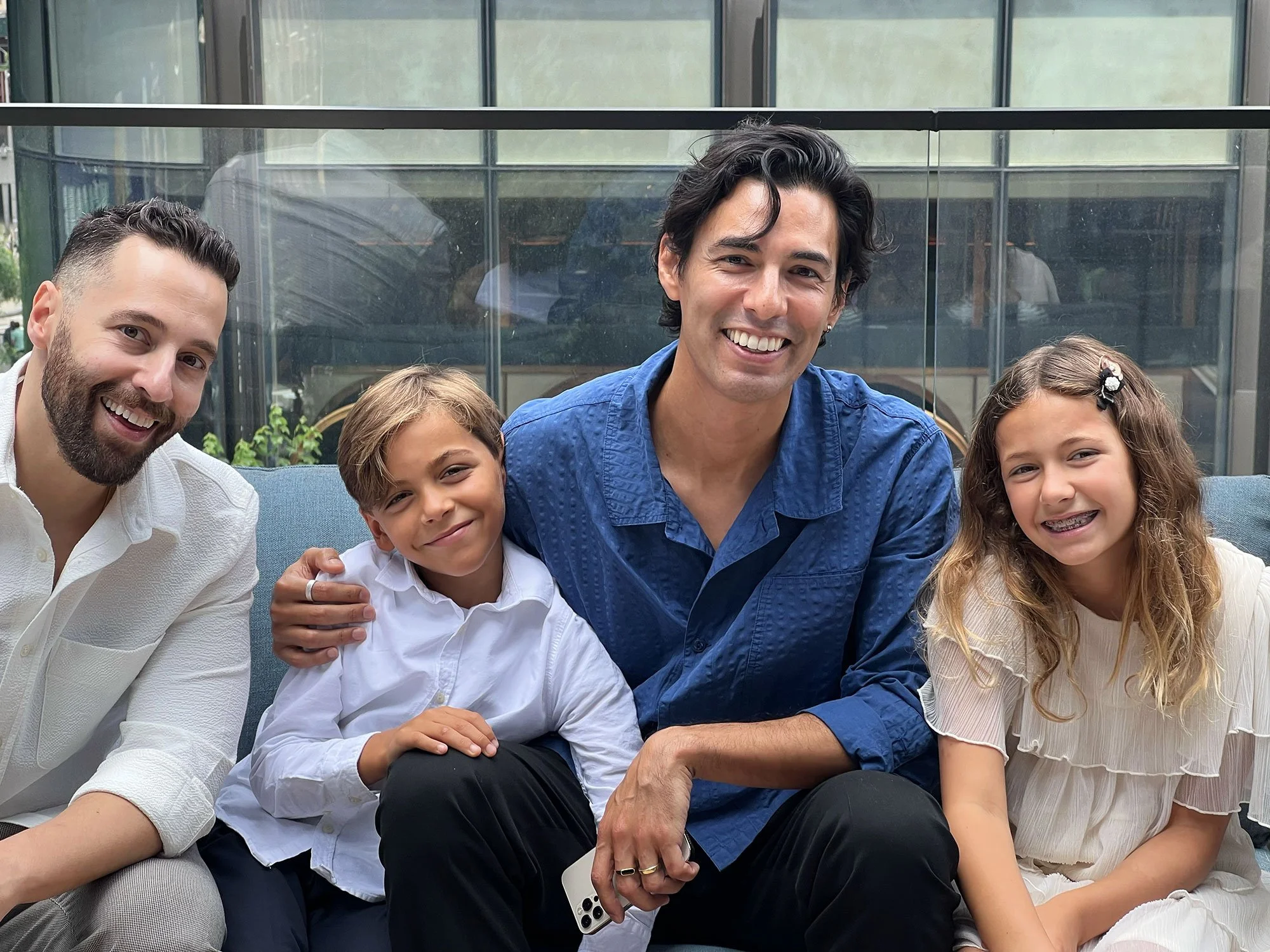Four people, two men and two children, sitting together and smiling outside on a balcony or patio with glass railing, enjoying a sunny day.