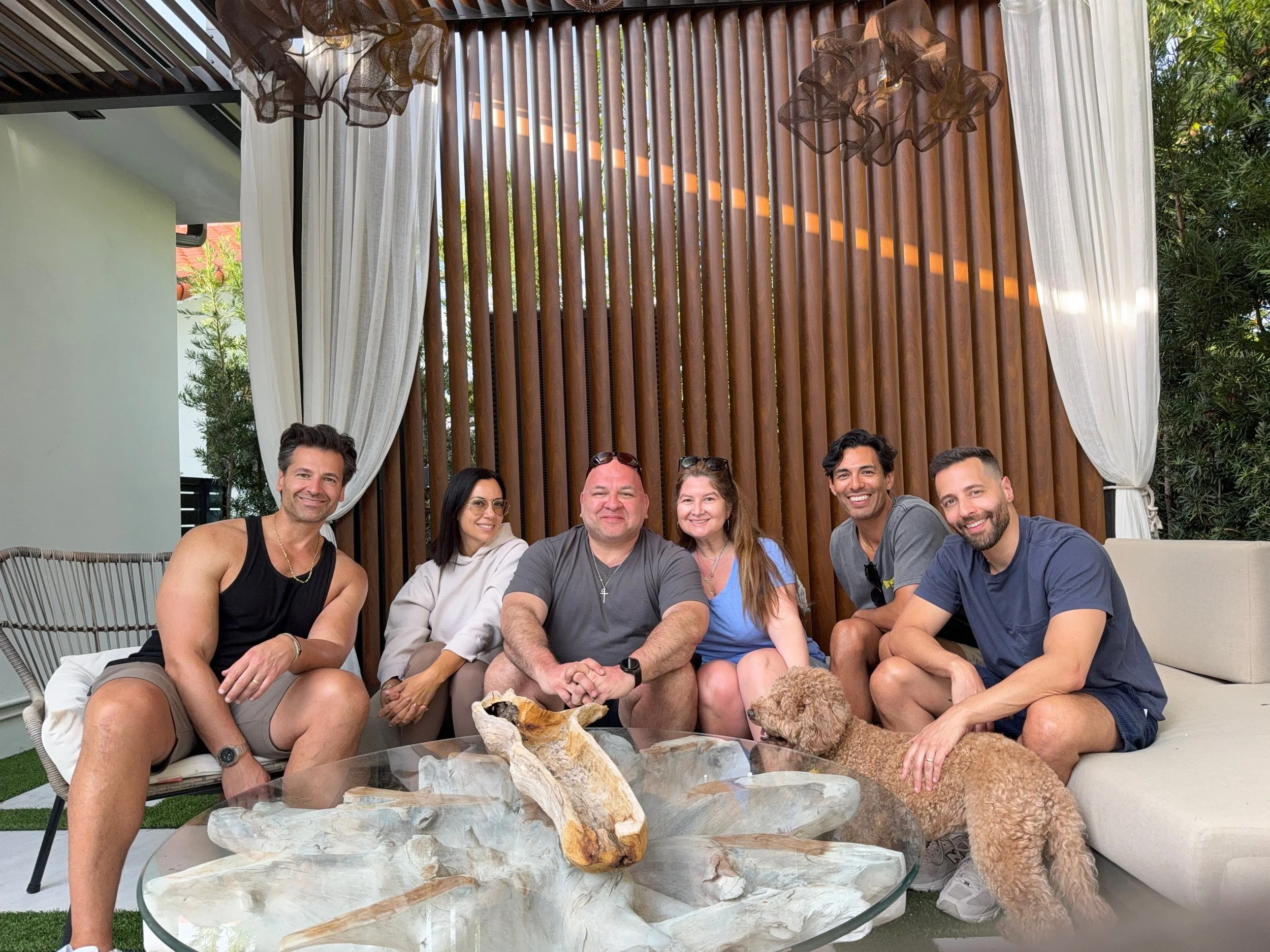 A group of six people sitting on outdoor furniture, smiling, with a small curly-haired dog near one person's feet, on a patio with a wooden slat wall and curtains in the background.