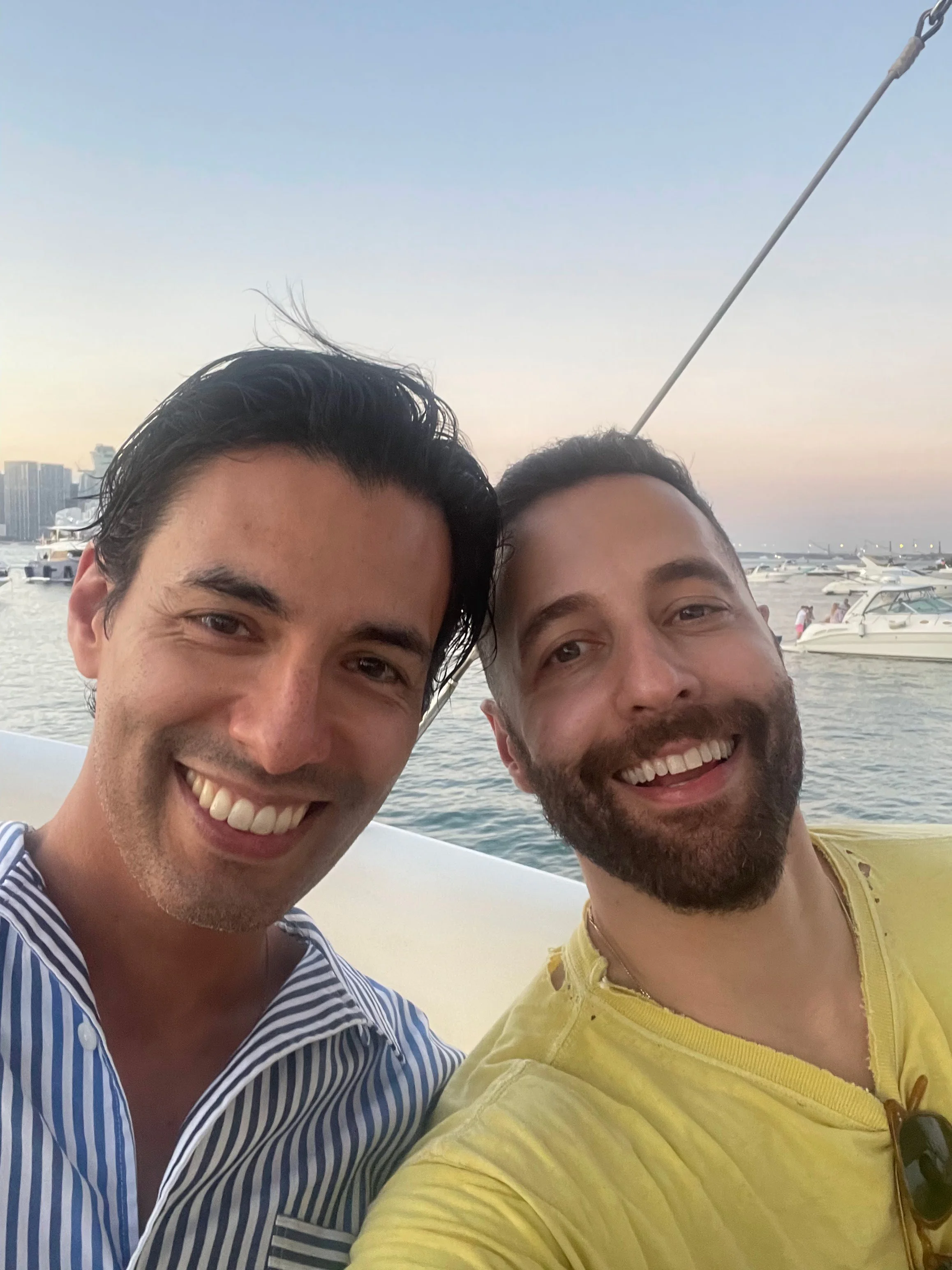 Two men smiling on a boat near a marina with yachts and city buildings in the background at sunset.