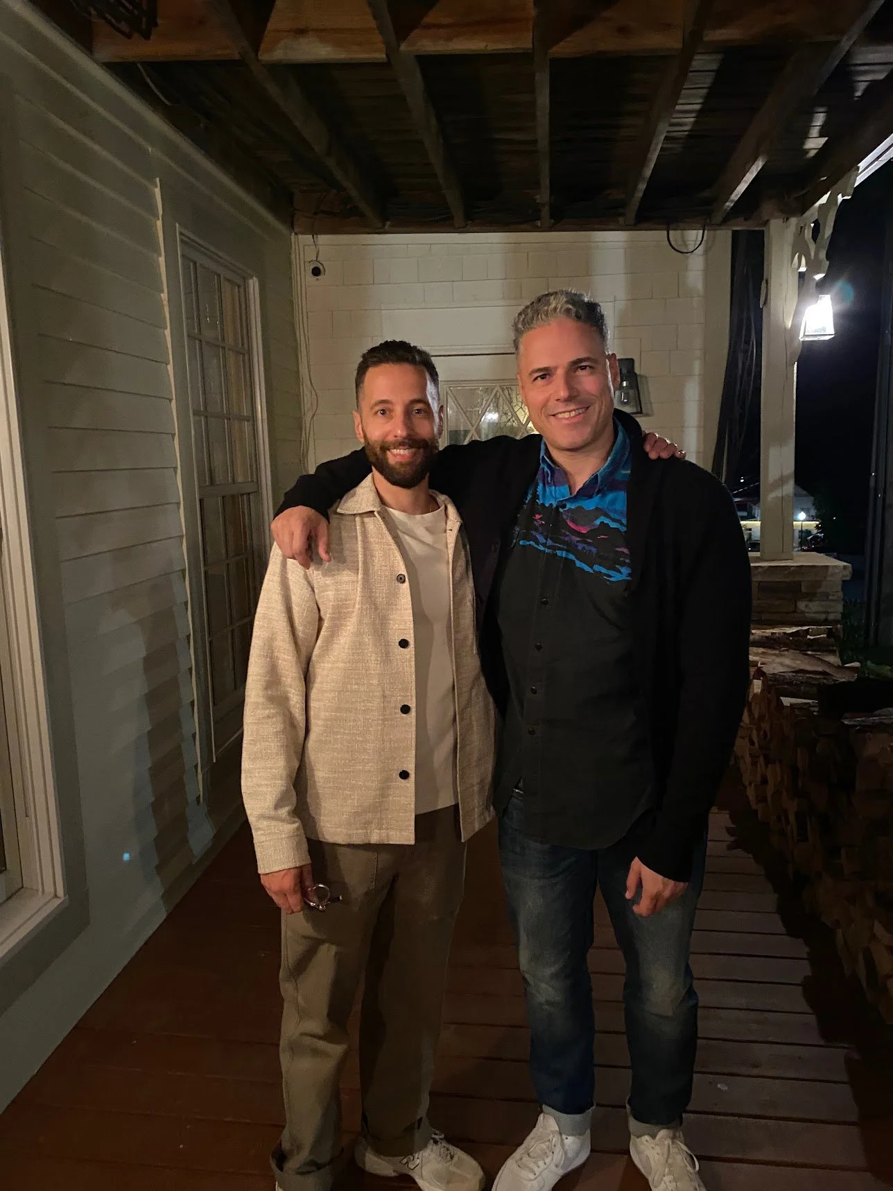 Two men standing together on a porch at night, smiling, with one man resting his arm on the other's shoulder.