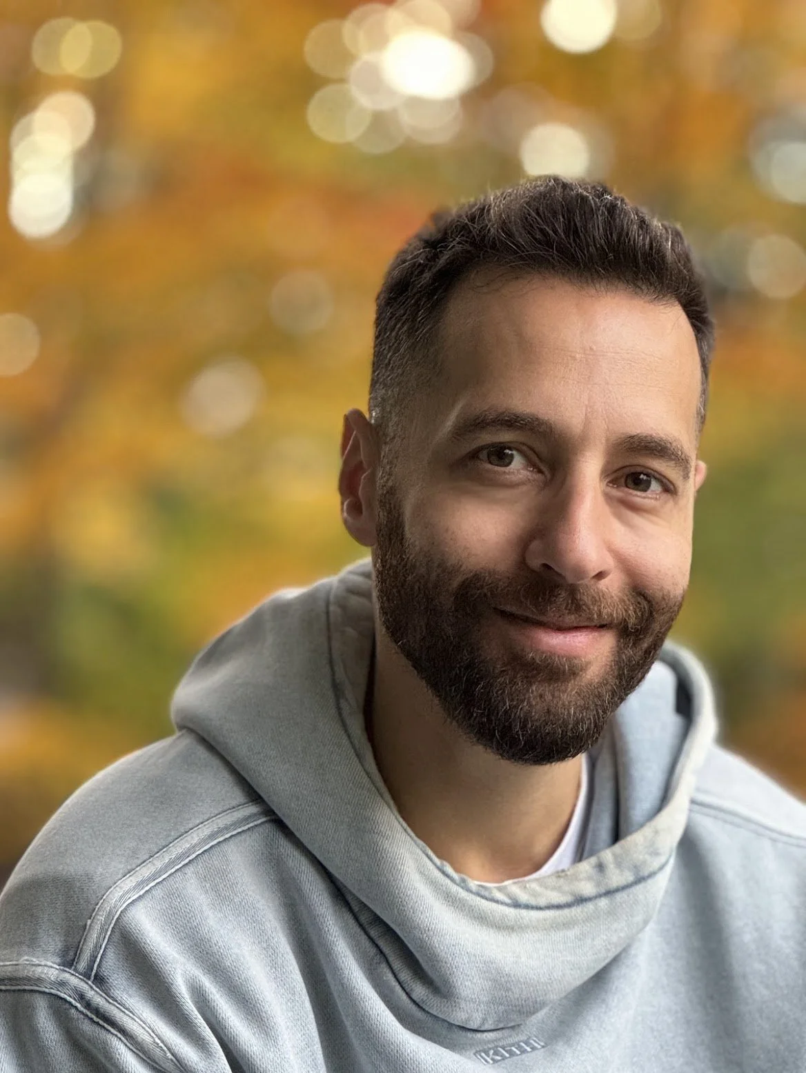 A man with dark hair, beard, wearing a gray hoodie, smiling outdoors with blurred autumn leaves in the background.