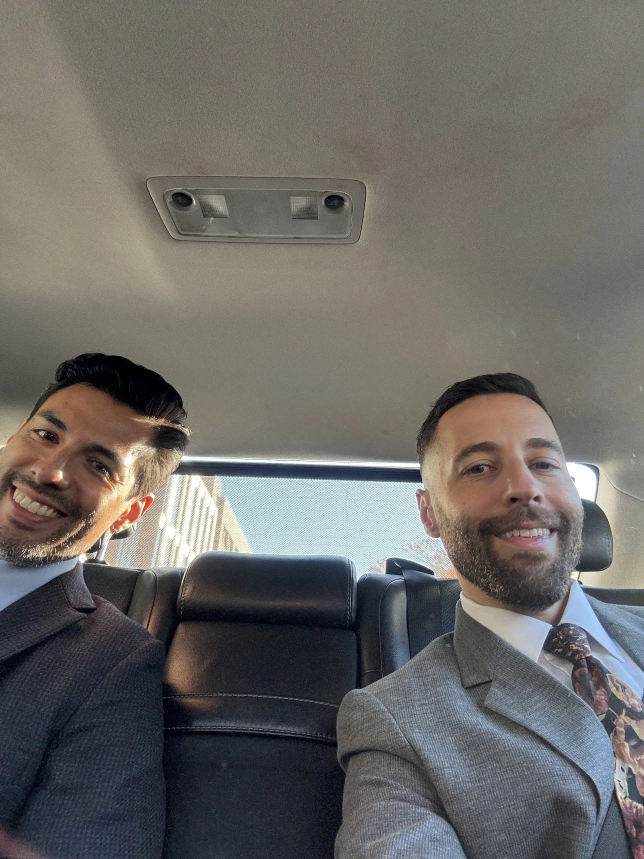 Two men in suits sitting in the backseat of a car, smiling for the photo.