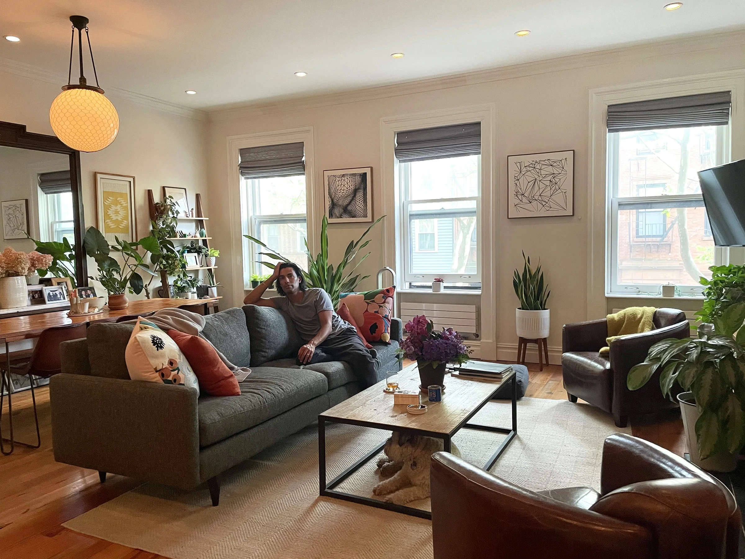 A living room with large windows, a gray sofa with colorful pillows, a wooden coffee table, and various potted plants. A person is sitting on the sofa, and a person is lying on the floor under the table. The room has hanging lights, framed artwork, and a bookshelf.