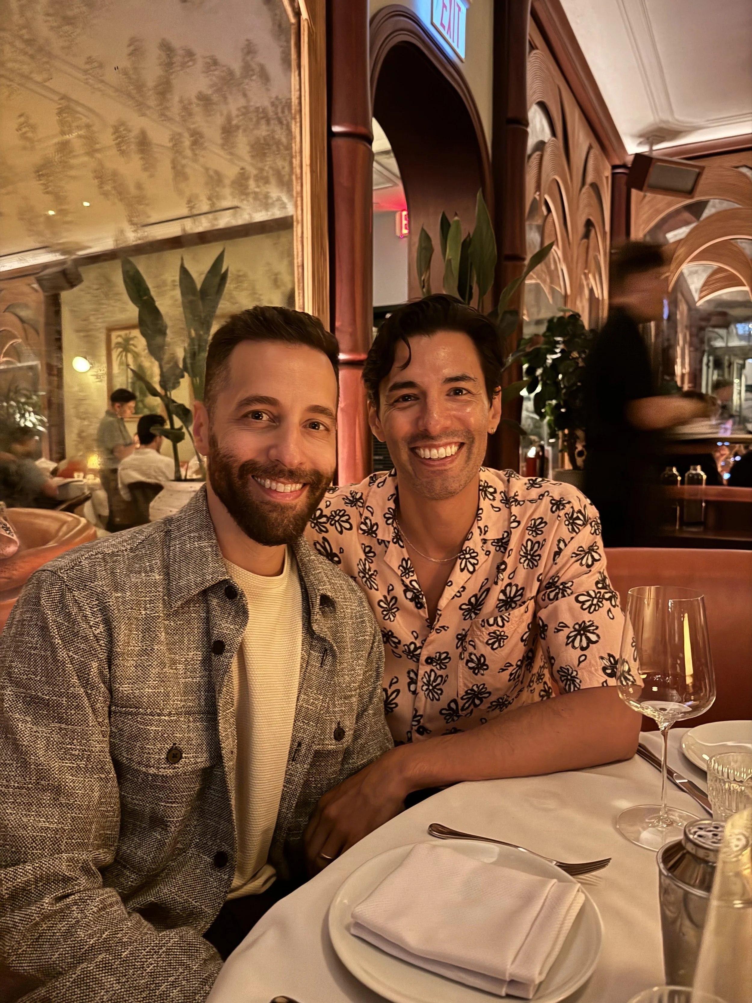 Two men sitting at a restaurant table, smiling at the camera, with a woman blurred in the background serving table and a large mirror on the wall behind them.